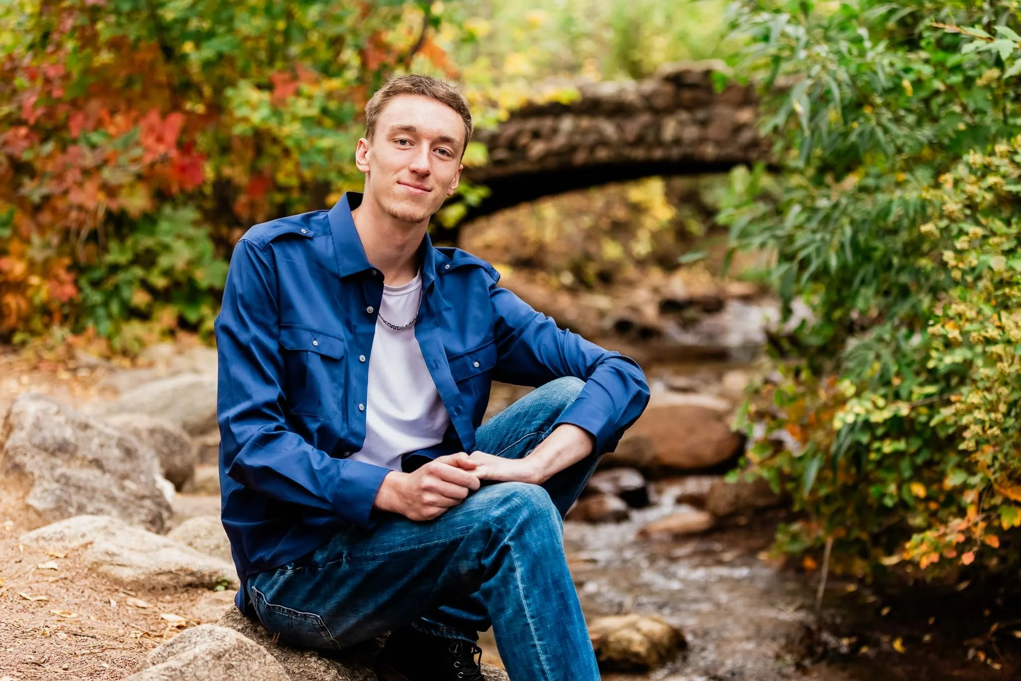 A young man sitting on rocks near a small stream in a forest with autumn-colored foliage and a small stone bridge in the background.