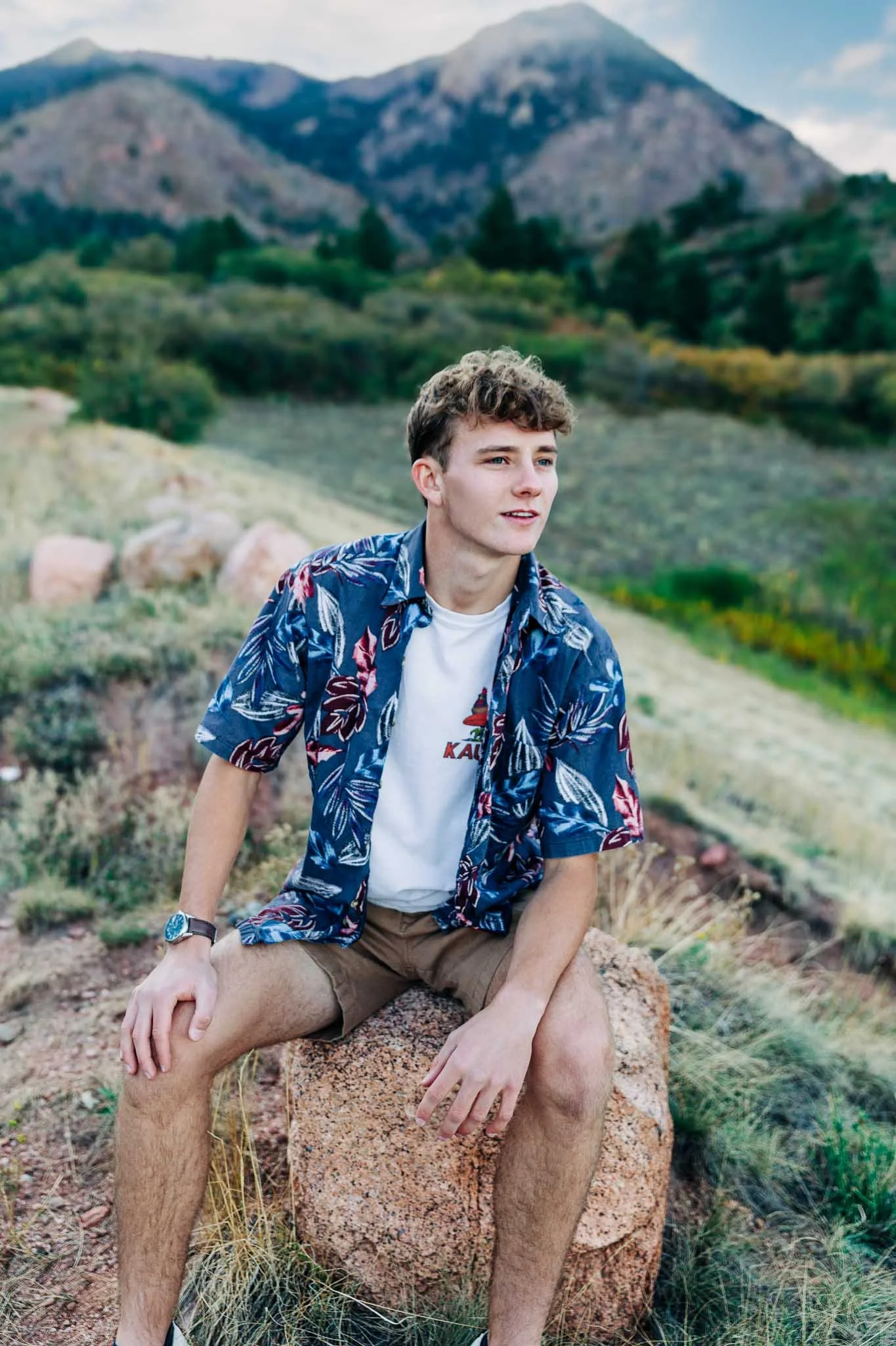 A young man with curly hair sitting on a rock in a mountainous landscape during daytime, wearing a blue floral Hawaiian shirt, a white T-shirt, and khaki shorts.