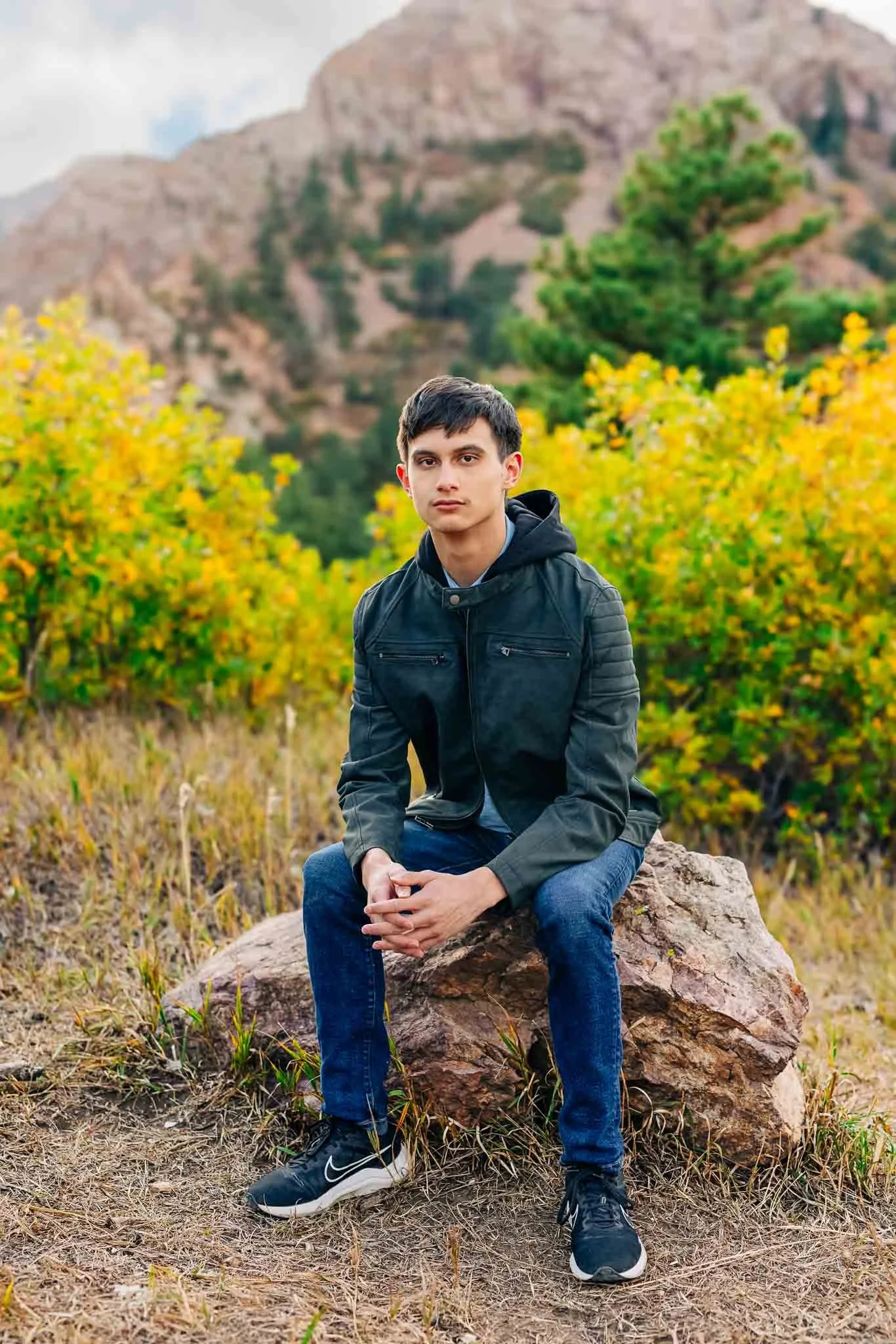 A young man in a black leather jacket and jeans sitting on a large rock in a natural landscape with green and yellow foliage and mountains in the background.
