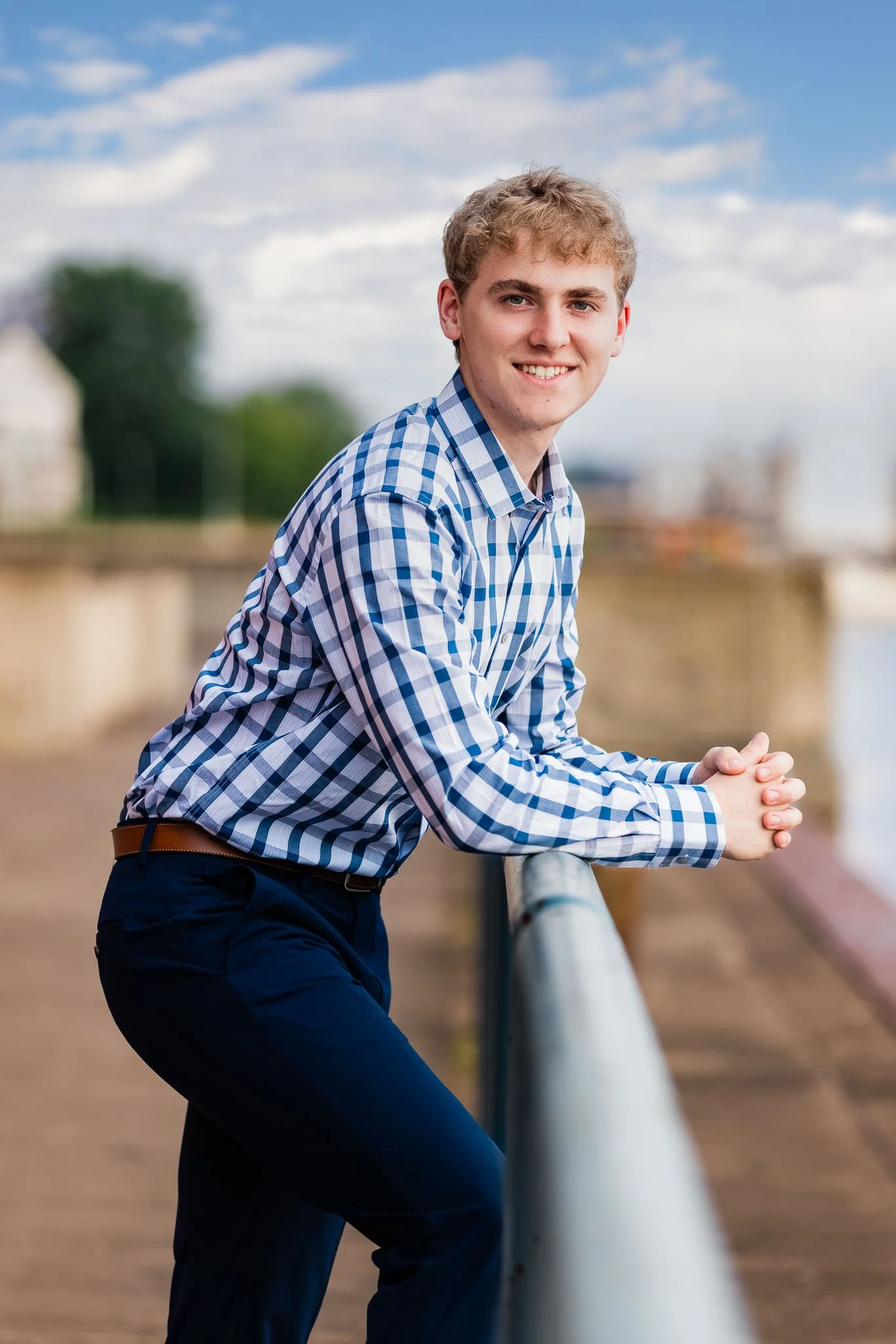 Young man in a checkered blue and white shirt leaning on a railing outdoors by a river, smiling at the camera with trees and a cloudy sky in the background.
