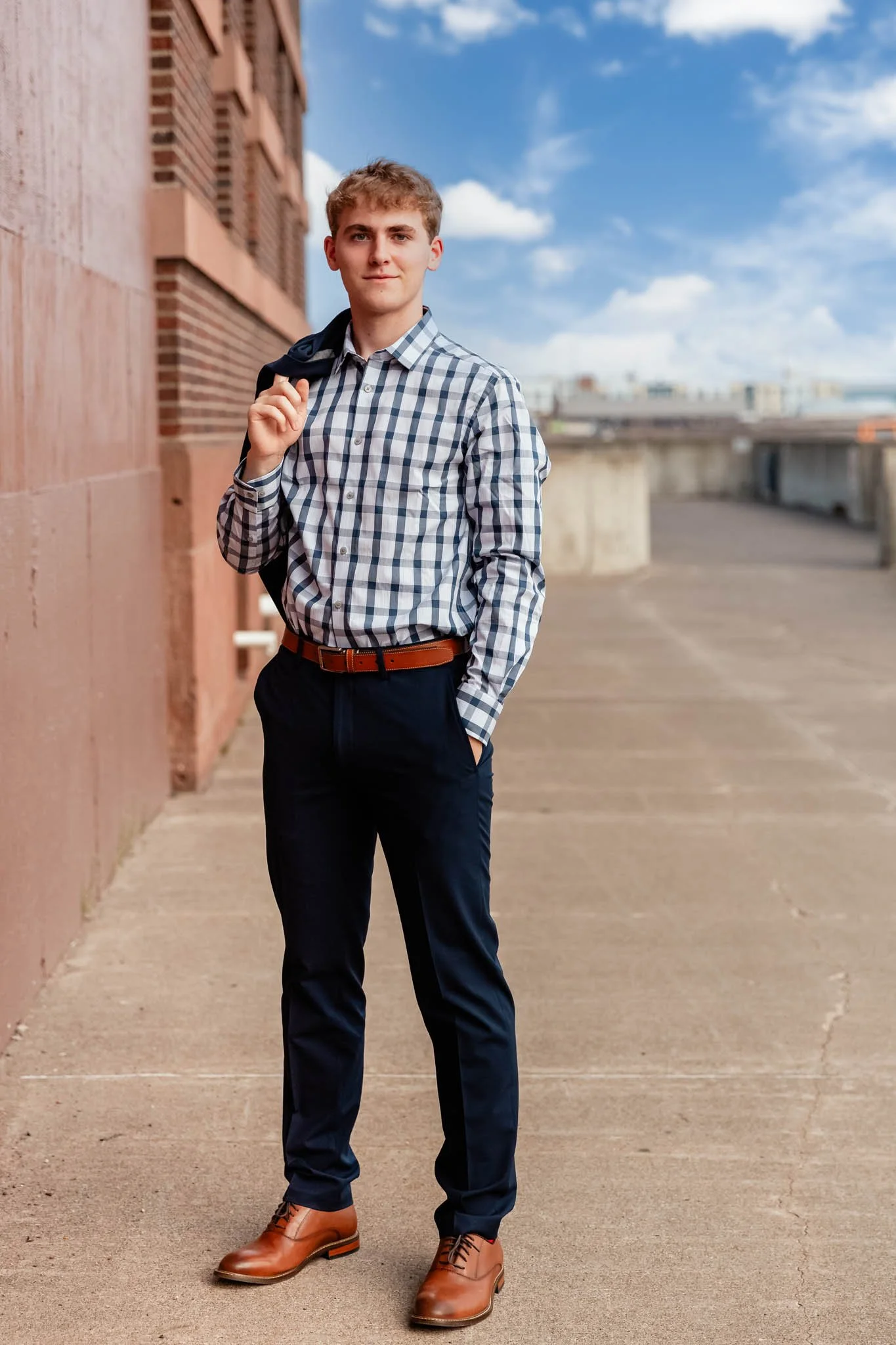 A young man standing on a city sidewalk wearing a checkered shirt, navy pants, and brown dress shoes, carrying a jacket over his shoulder, with a brick building and a cloudy sky in the background.