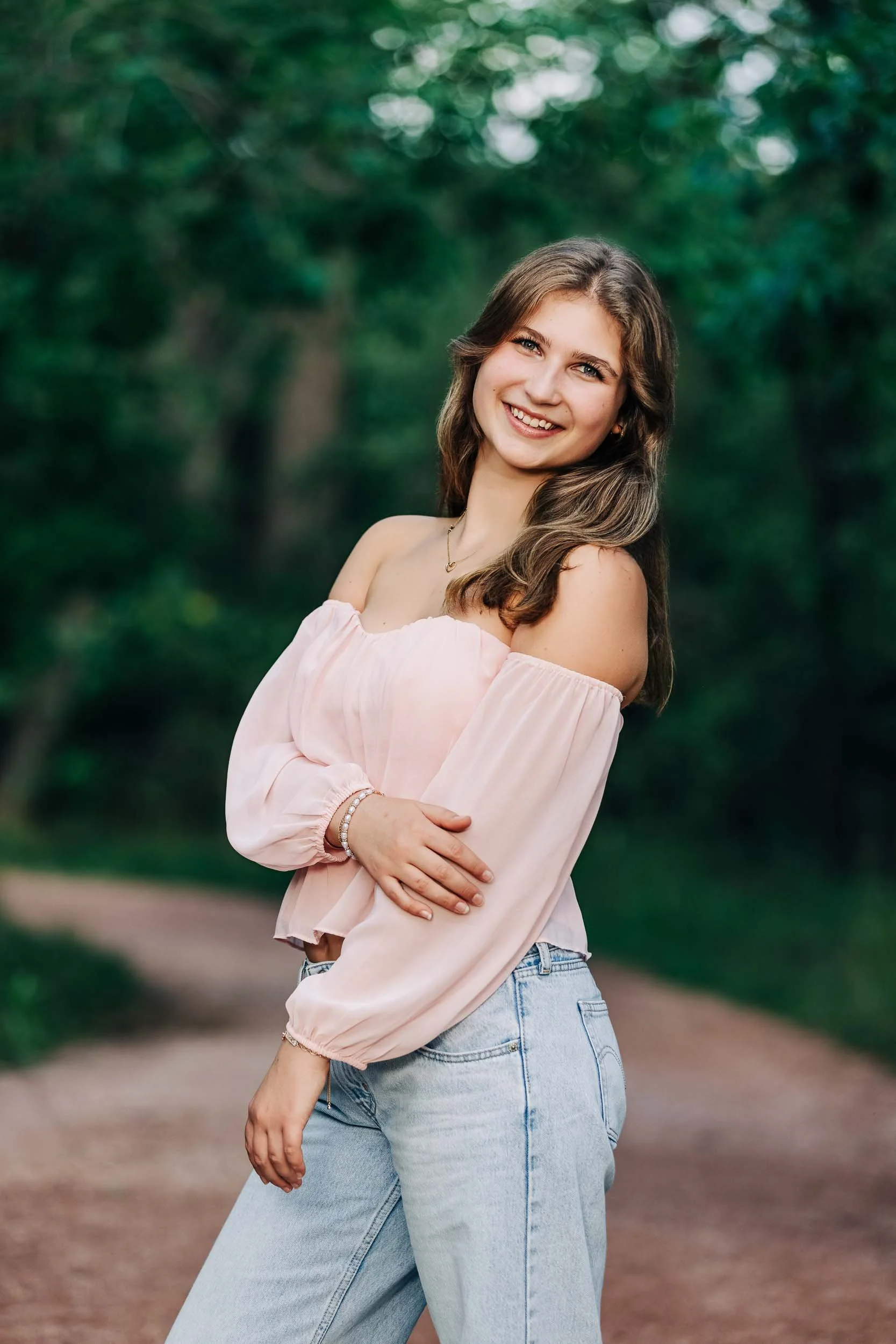 A high school girl with brown hair smiling outdoors in a pink off-shoulder top and light blue jeans.