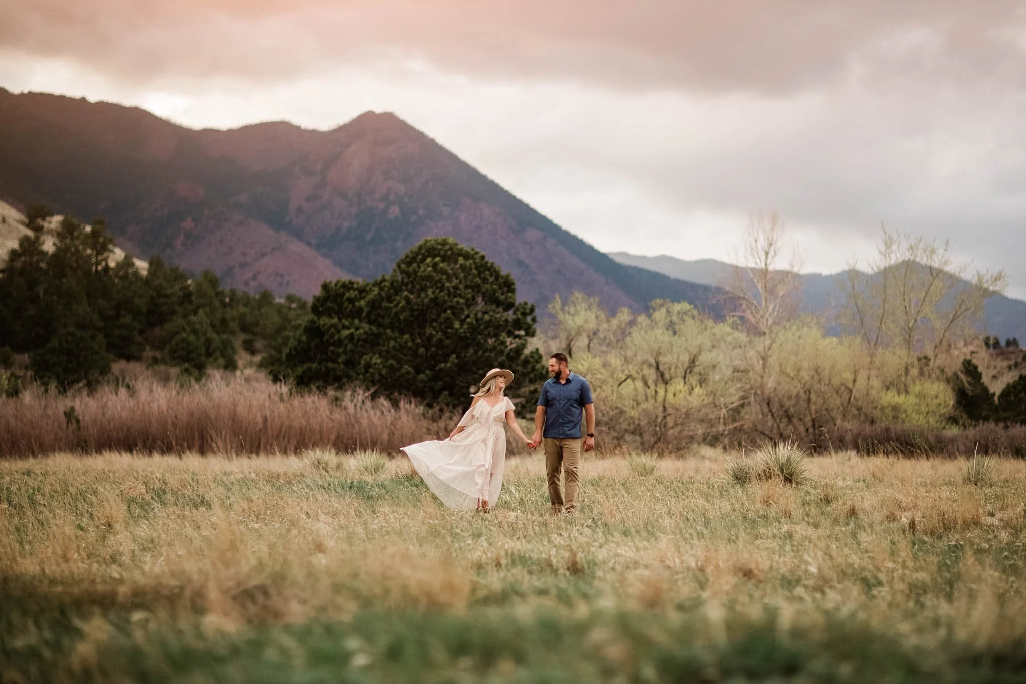 A couple holding hands and walking through a grassy field with mountains in the background, during a cloudy day.
