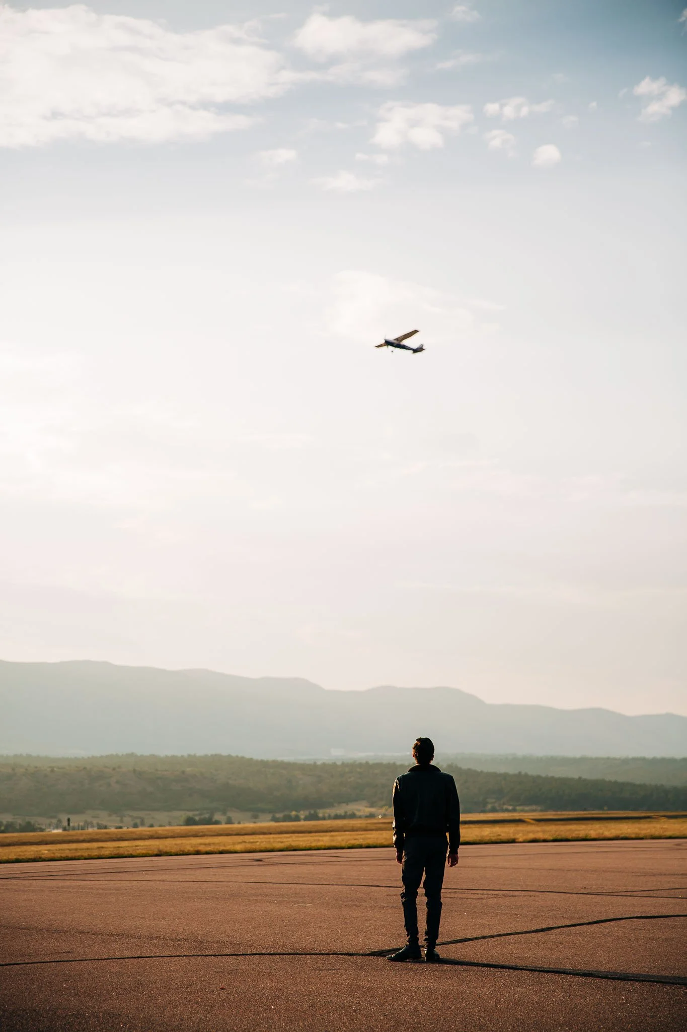 A man stands on an empty tarmac watching a small airplane flying in a sky with scattered clouds and mountains in the background.