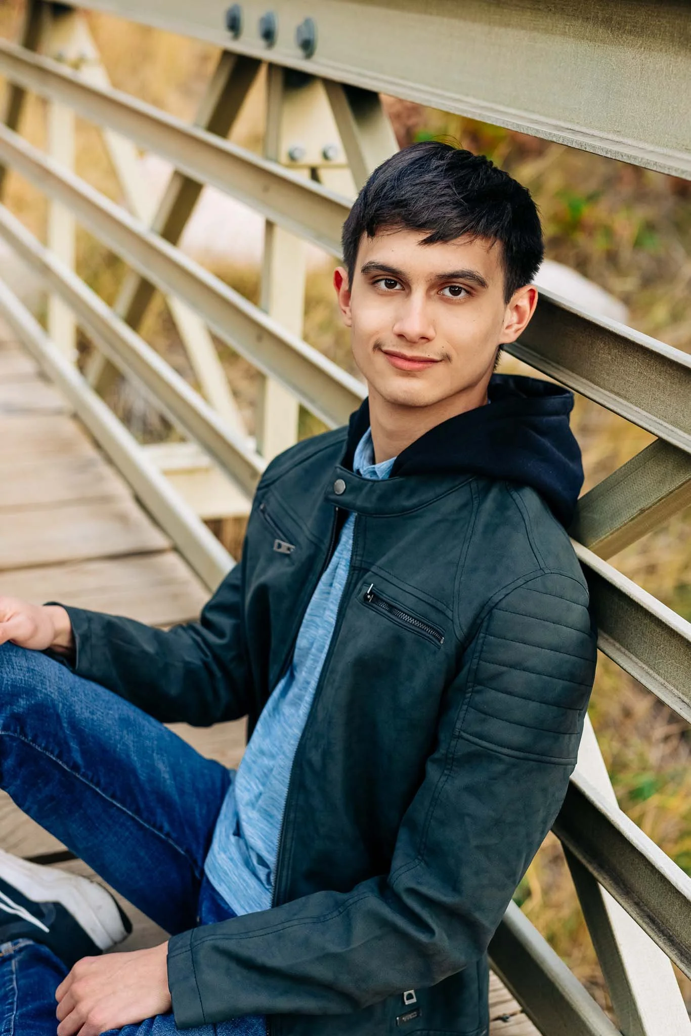 A young man with dark hair and light skin sitting on a wooden walkway, leaning against a railing, wearing a black jacket and blue jeans, outdoors with a blurred background of autumn foliage.