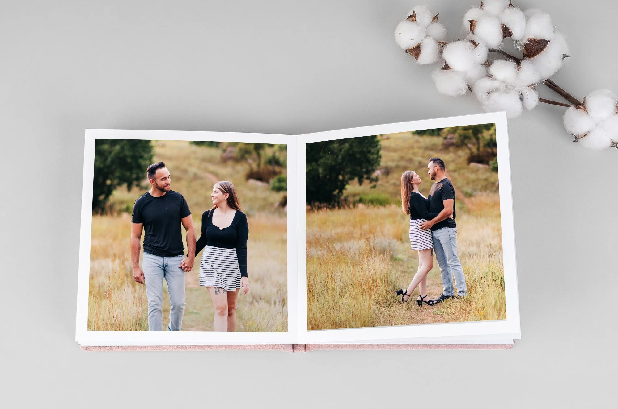 Open photo album with two pictures of a couple walking and embracing outdoors in a grassy field, with a cotton branch in the top right corner.