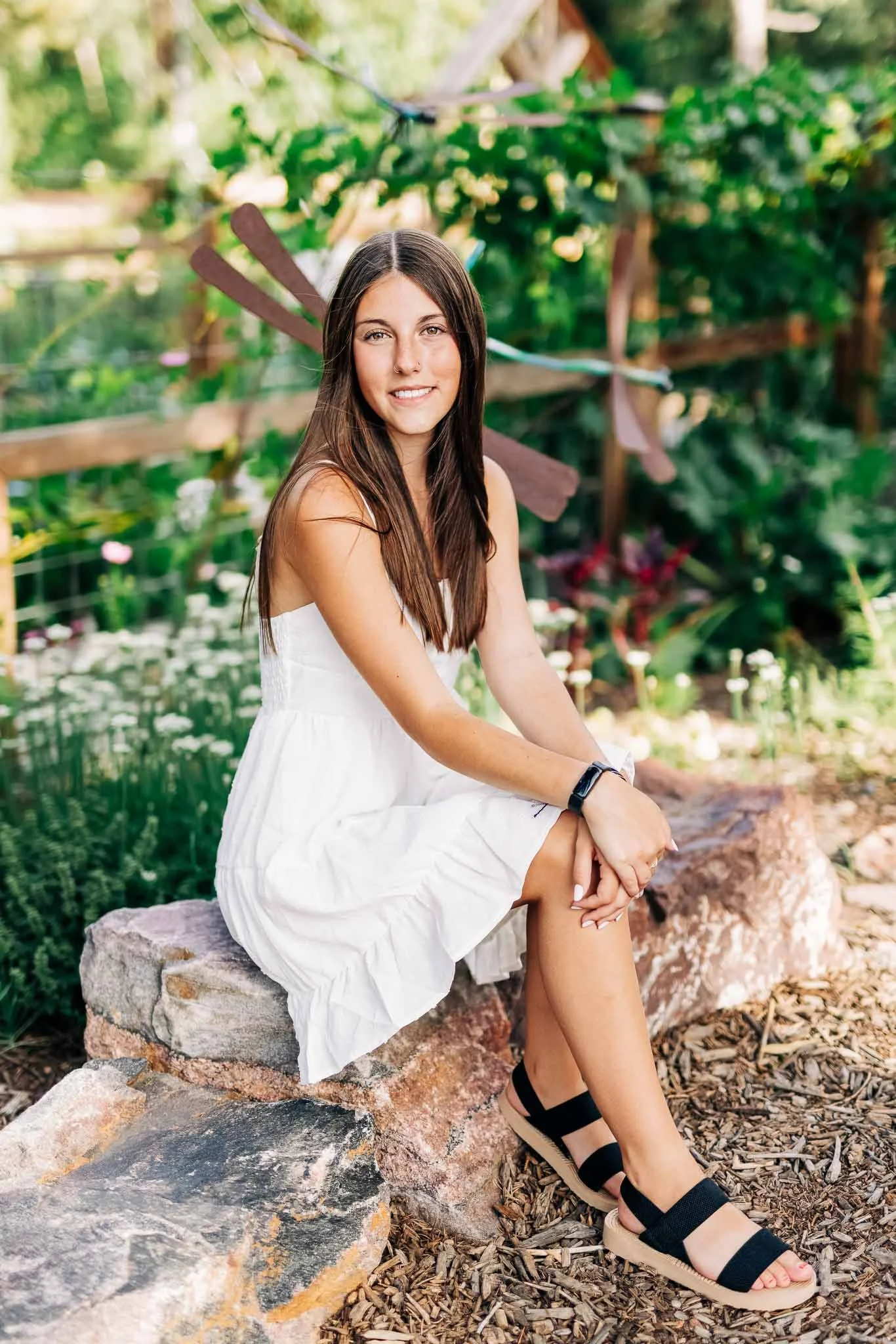 A young woman with long brown hair, wearing a white sundress and black sandals, sitting on a rock in a garden with lush greenery and a wooden post in the background.