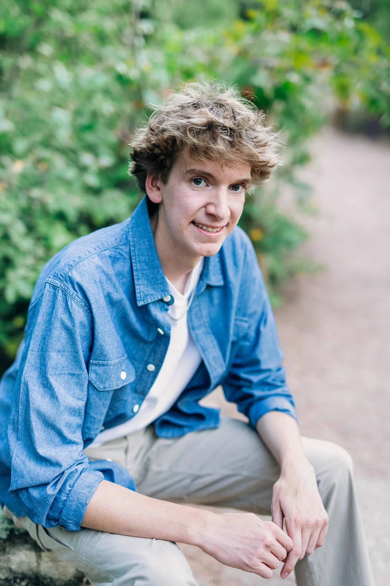 A young man with curly blond hair and blue eyes, wearing a blue denim shirt over a white t-shirt, sitting outdoors on a dirt path surrounded by green foliage, smiling at the camera.