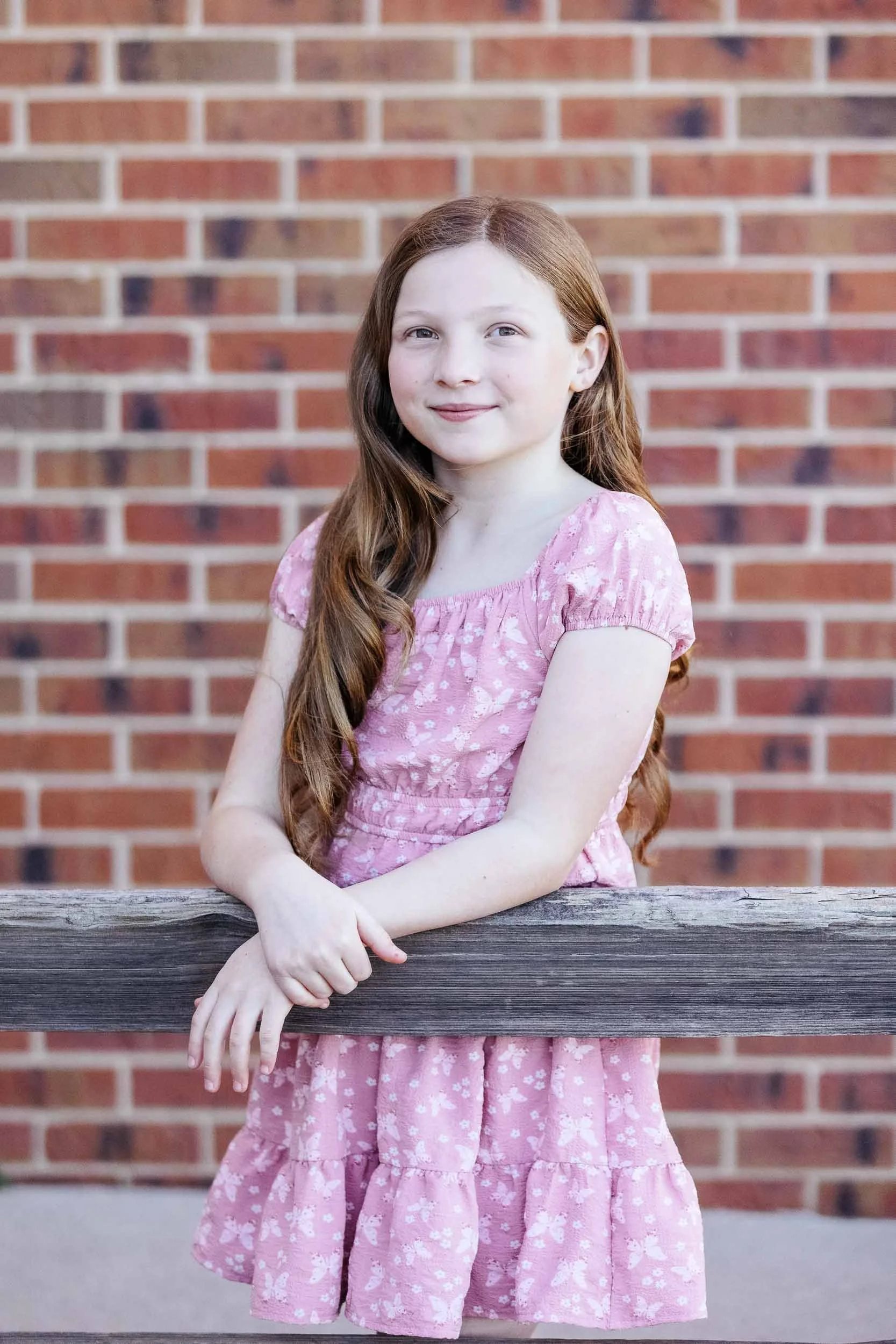 A young girl with long red hair, wearing a pink dress with a white floral pattern, standing outdoors in front of a brick wall, resting her forearms on a wooden railing, smiling softly.
