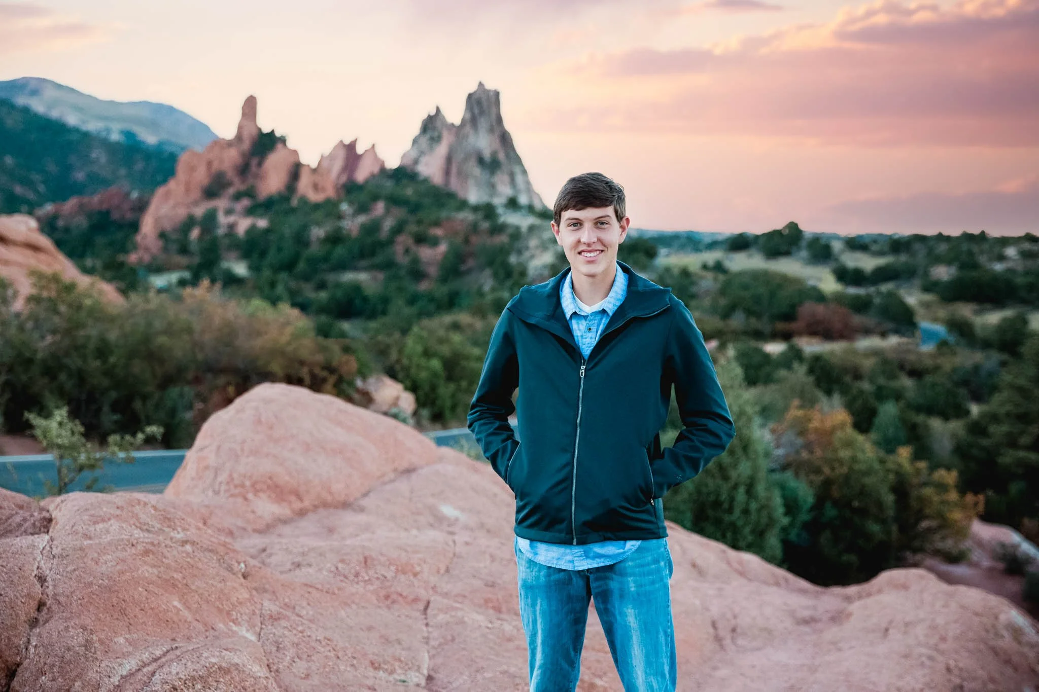 A young man stands on reddish rocks with mountains and trees in the background during sunset, smiling at the camera.