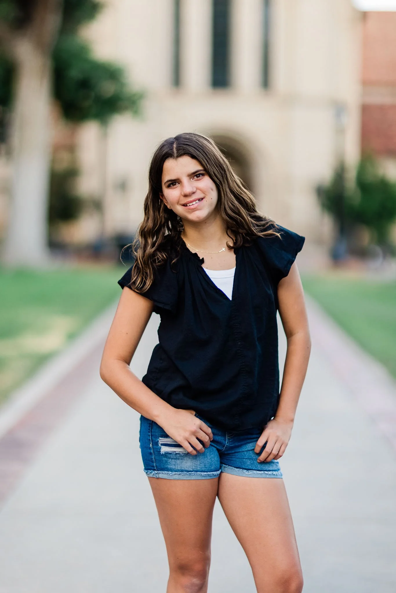 Young woman standing on a sidewalk in front of a building, wearing a black top and denim shorts, smiling at the camera.
