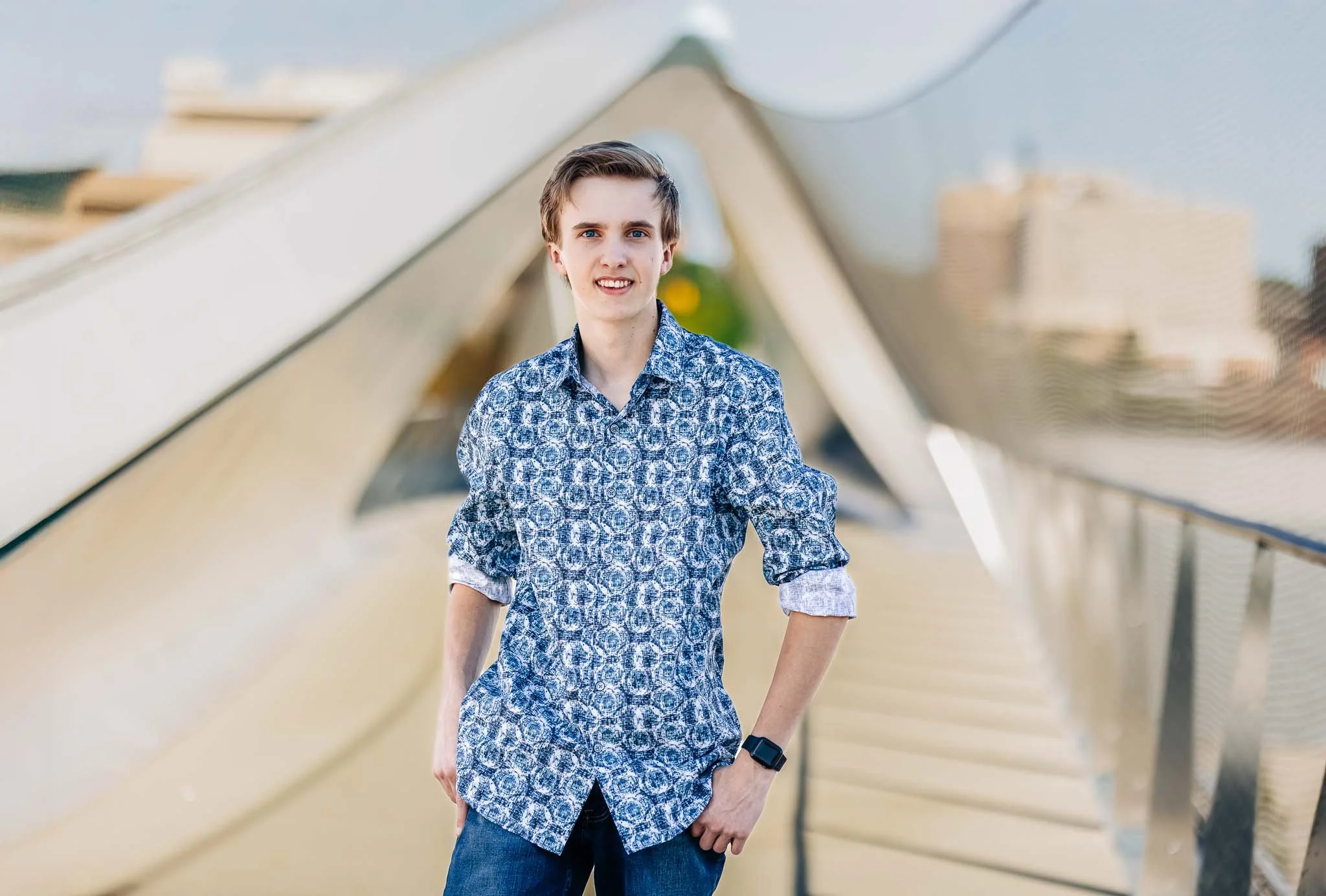 A young man with brown hair and blue eyes wearing a blue patterned shirt with rolled-up sleeves and dark jeans, standing outdoors on a bridge or walkway with metallic railings, smiling at the camera.