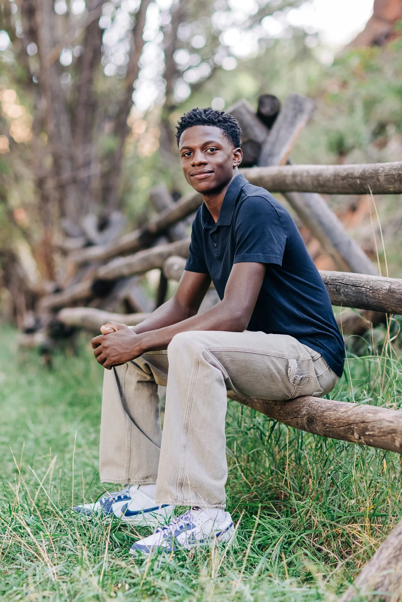 A high school senior guy with dark curly hair sitting on a wooden fence in a natural outdoor setting, wearing a black polo shirt, beige pants, and white sneakers with blue accents.