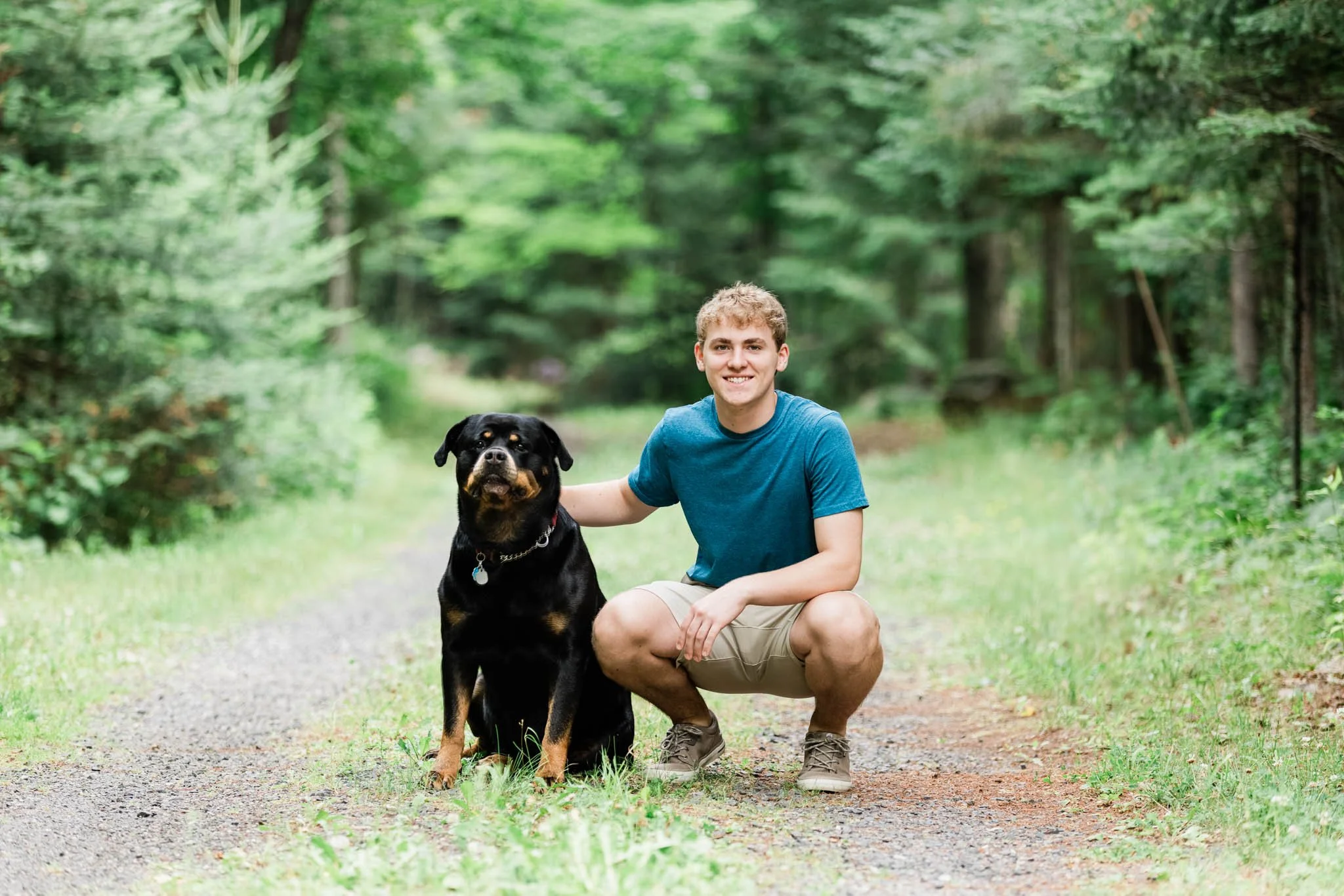 A young man crouching next to a Rottweiler dog on a forest trail during daytime.