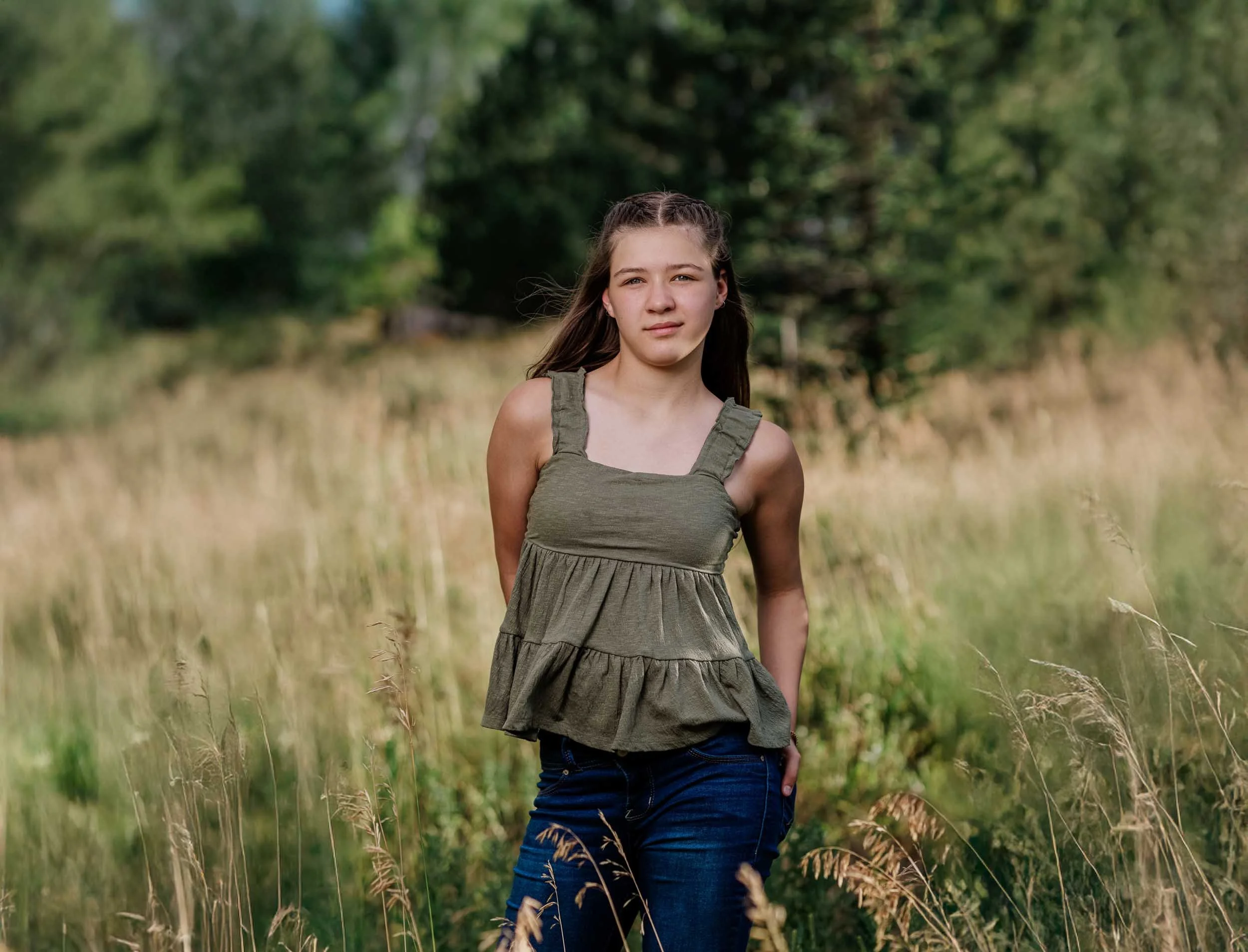 A young girl standing in a field of tall grass with trees in the background, wearing a green sleeveless top and dark jeans.