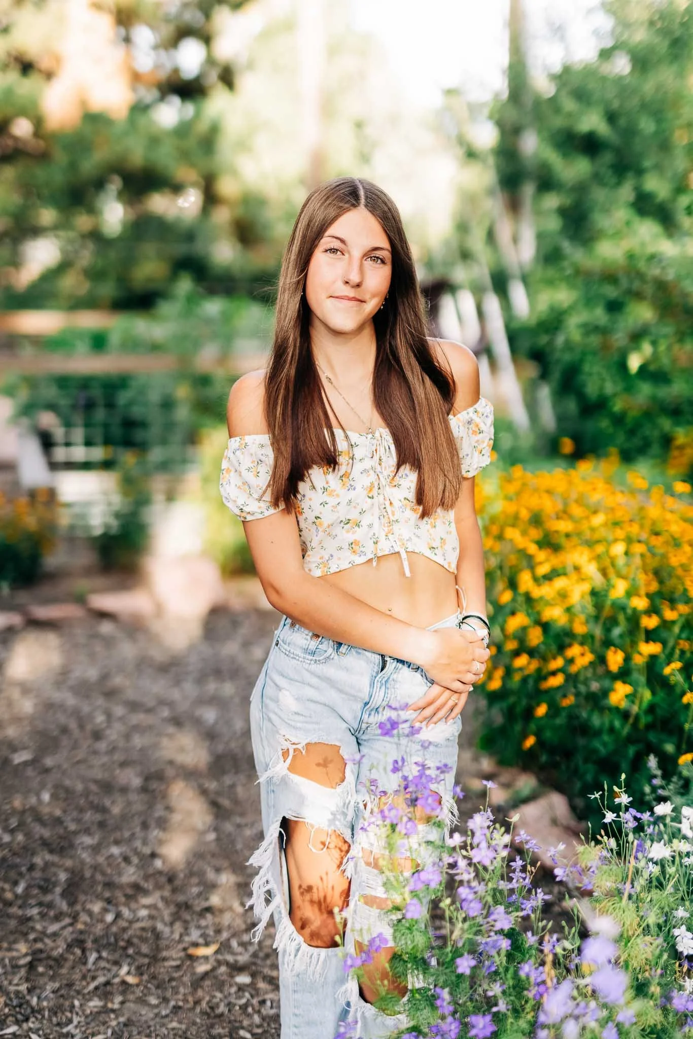 Young woman with long brown hair standing in a garden filled with colorful flowers, wearing a floral off-shoulder crop top and ripped jeans.