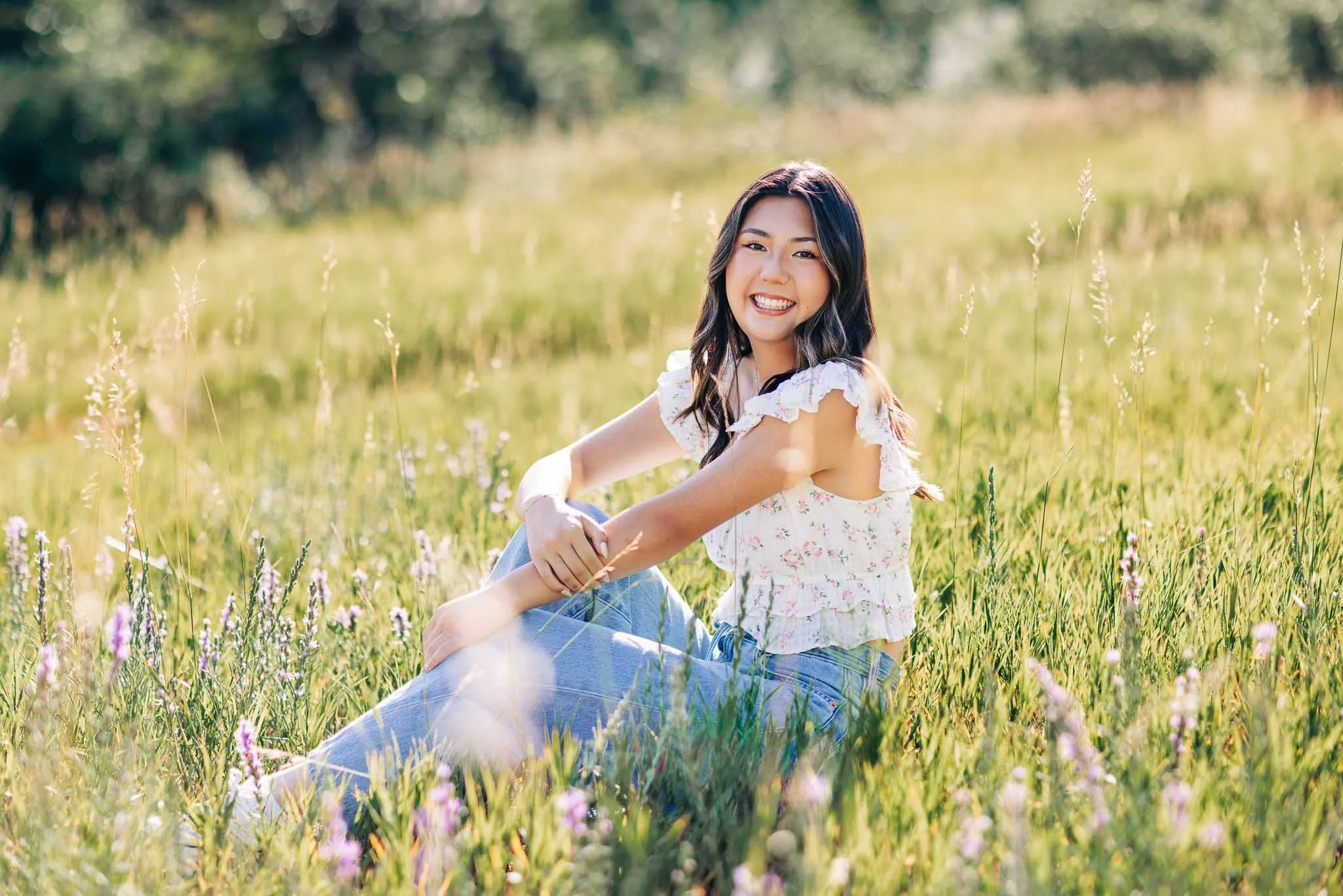 A young woman sitting in a grassy field with purple flowers, smiling and winking at the camera.