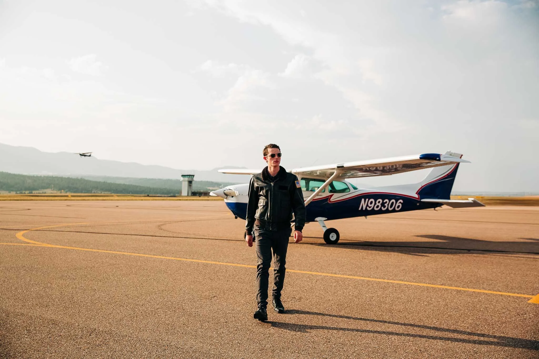 A man walking on an airport tarmac in front of a small private airplane, with another aircraft flying in the background.