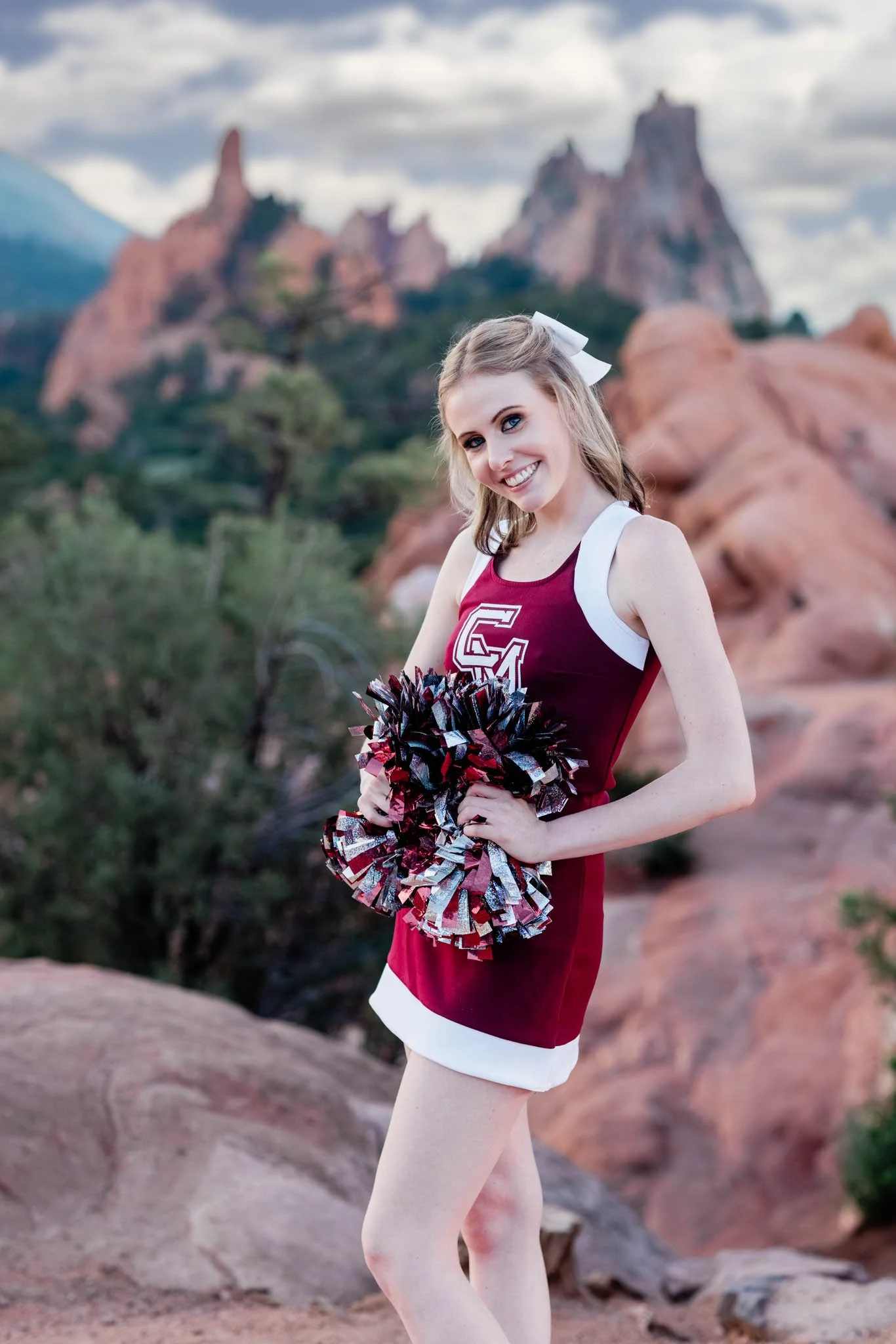 A young woman in a cheerleader outfit smiling with pom-poms outdoors against a rocky mountain landscape.