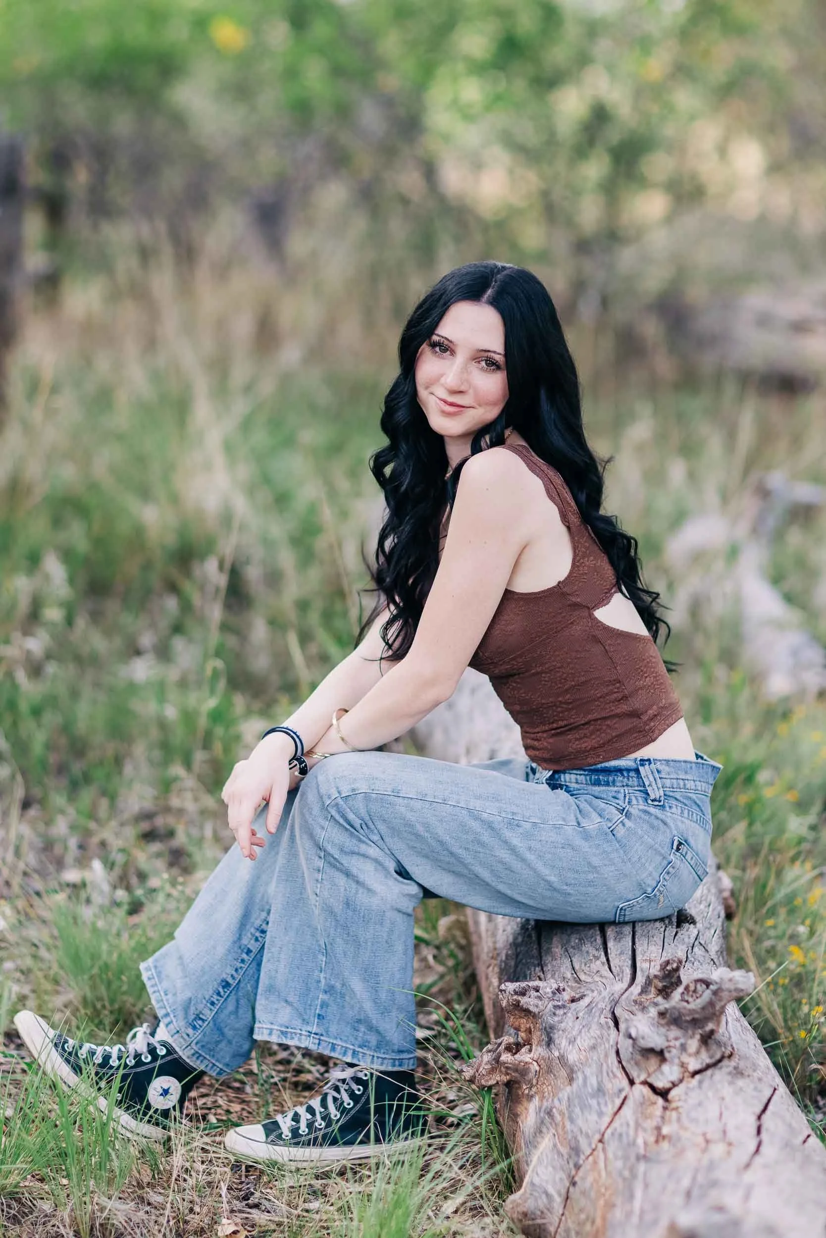 A high school senior girl with long black hair, wearing a brown tank top and ripped jeans, sitting on a log in an outdoor wooded area with green foliage. She is smiling at the camera.