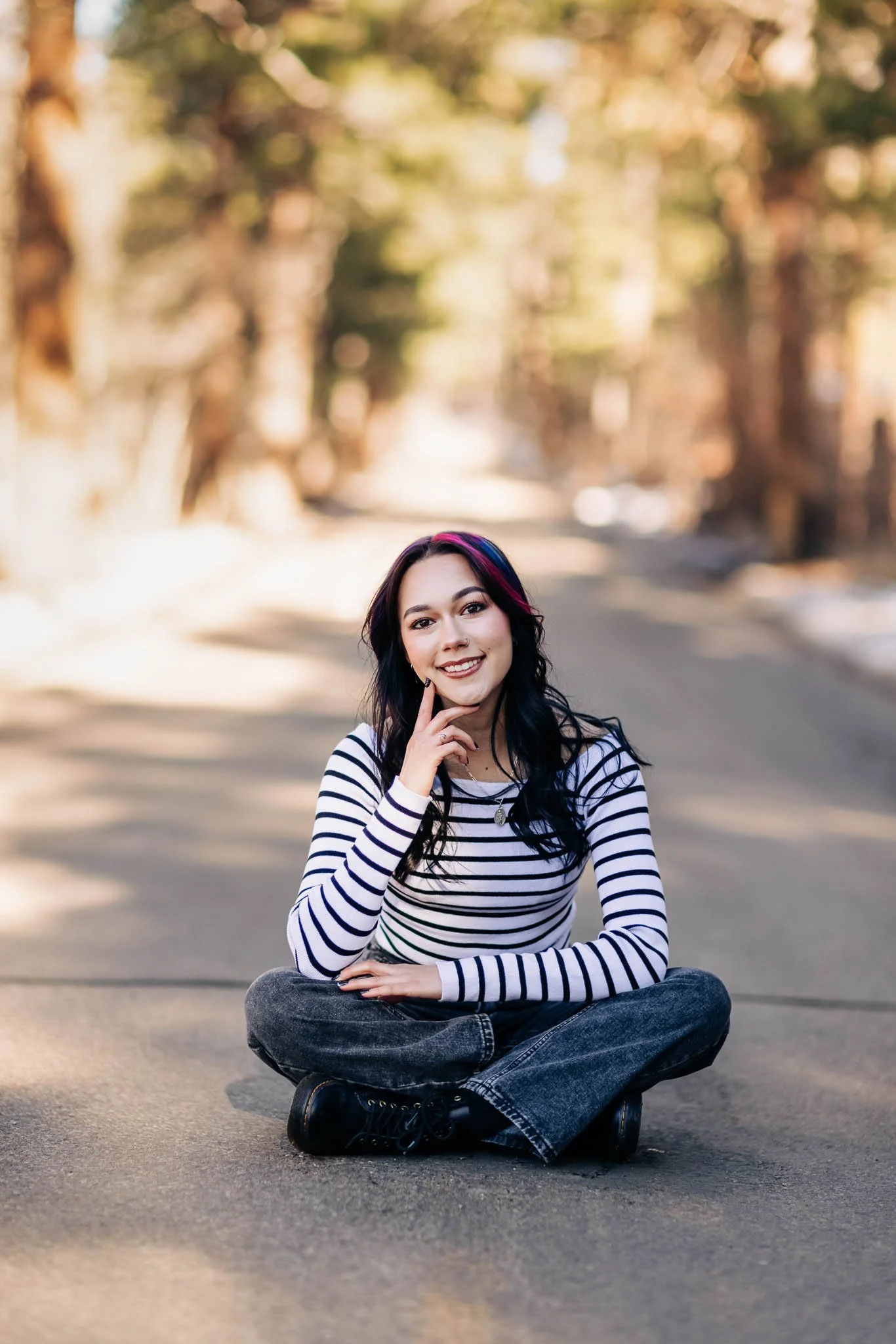 A young woman with dark hair and pink streaks, sitting cross-legged on a paved outdoor path, smiling at the camera with her hand resting on her chin. She is wearing a striped shirt and jeans, surrounded by a blurred background of trees and sunlight.