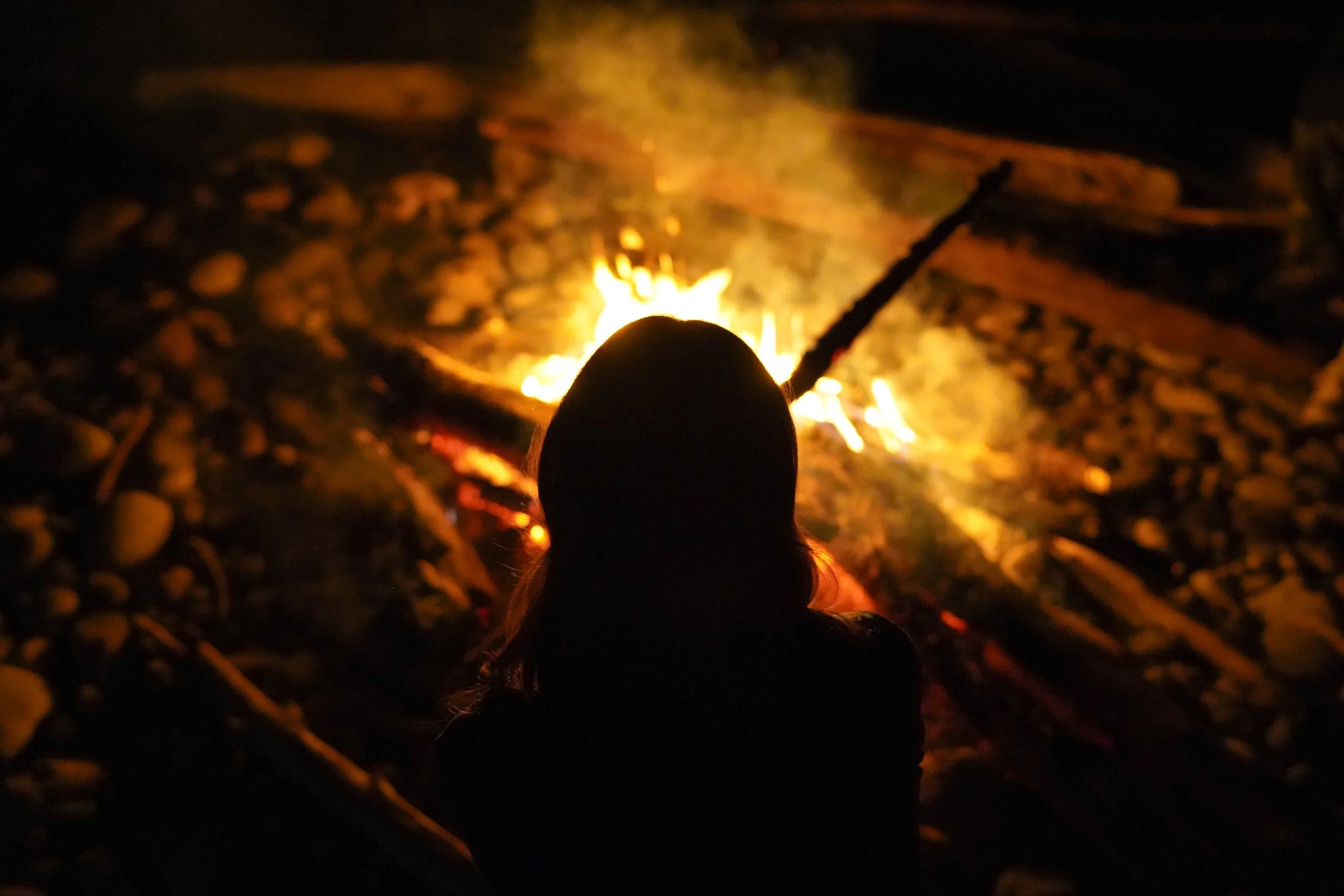 Silhouette of a person with long hair sitting by a campfire at night on a rocky surface.