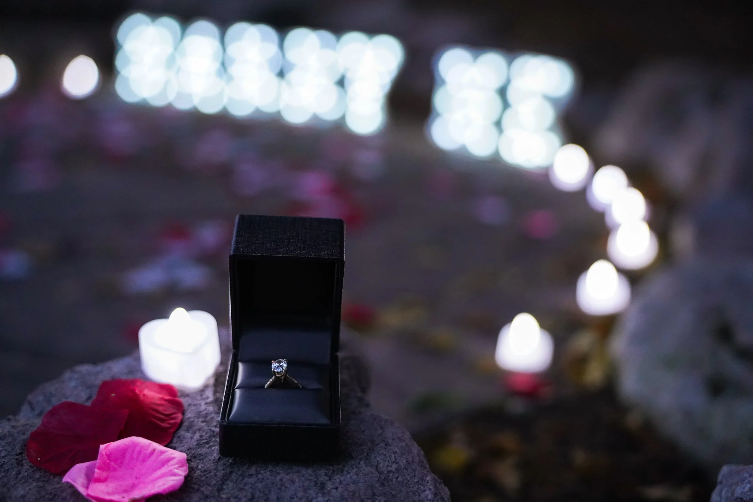 A diamond engagement ring in a black box, surrounded by white candle lights and flower petals on a rock at night.