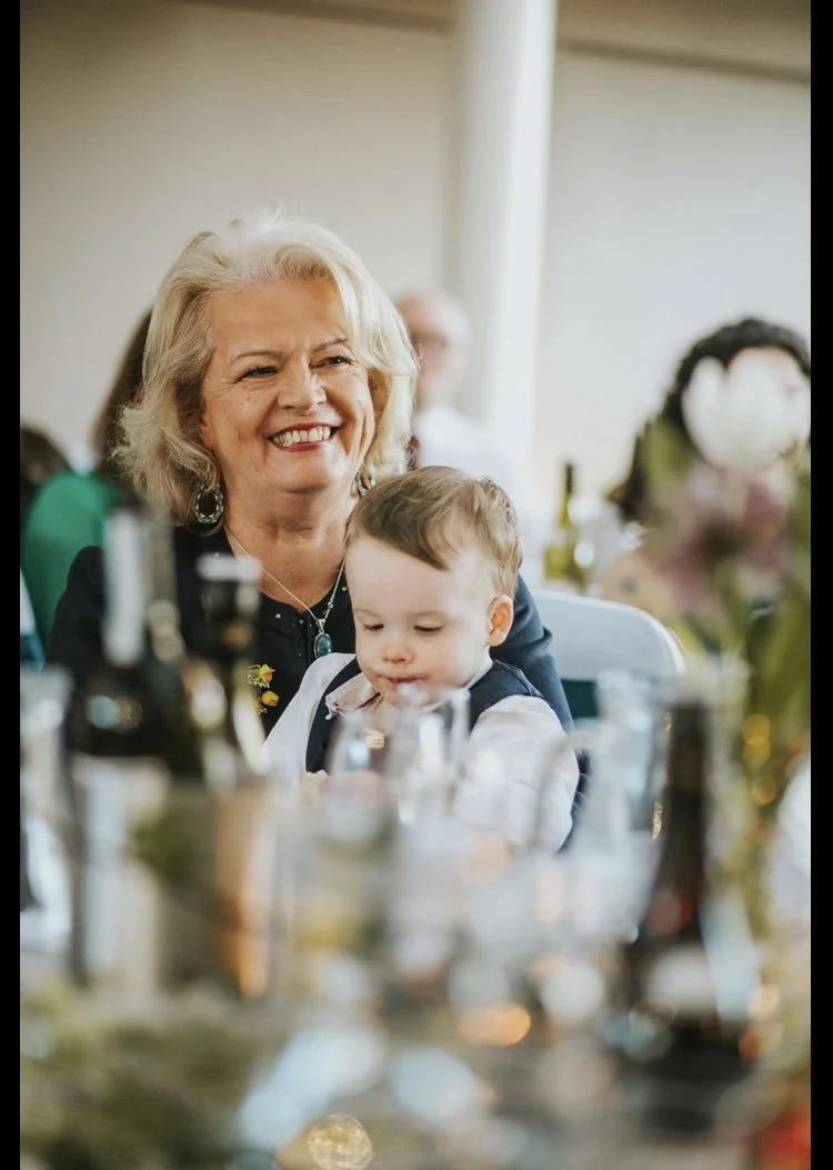 GILL BARHAM smiling while sitting at a table with a young child in her lap, in a social gathering setting.