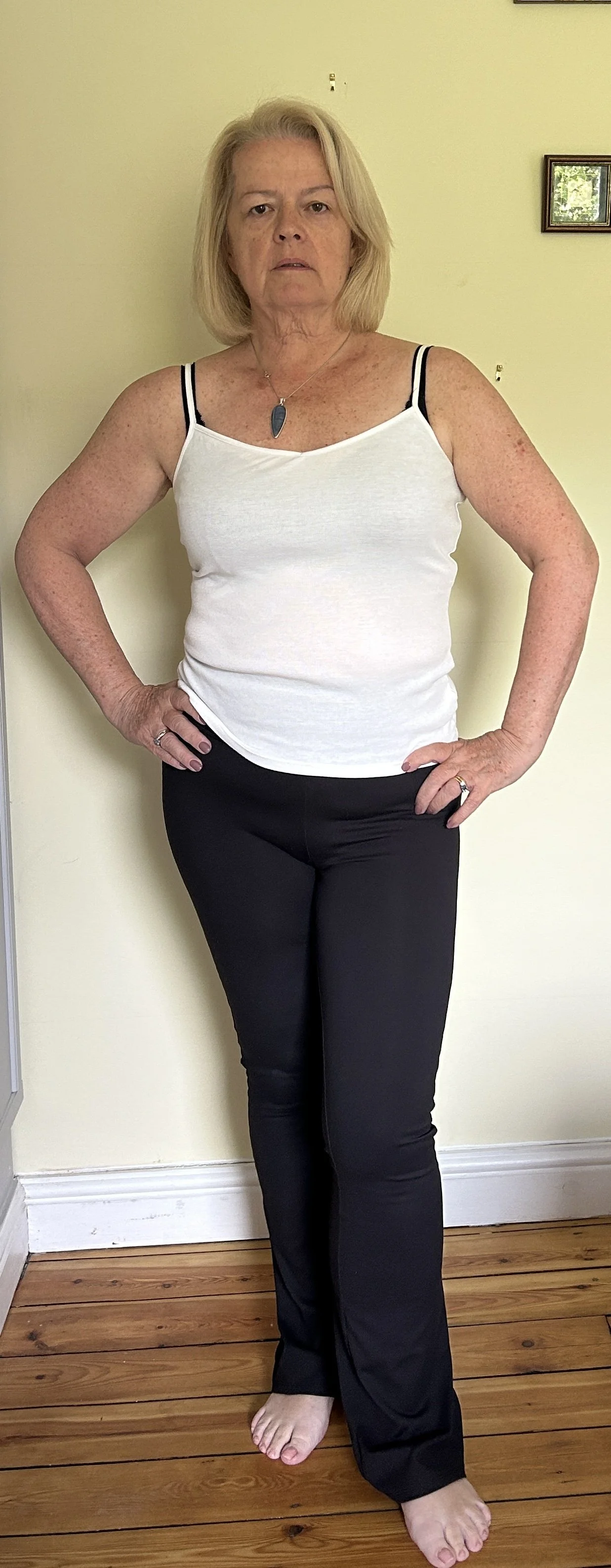 GILL BARHAM with shoulder-length blonde hair stands with her hands on her hips against a plain yellow wall, wearing a white tank top with black straps and black pants, and barefoot on a wooden floor.