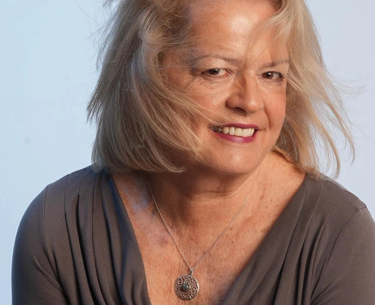 A close-up portrait of an GILL BARHAM with blonde hair, smiling, wearing a grey top and a silver pendant necklace.