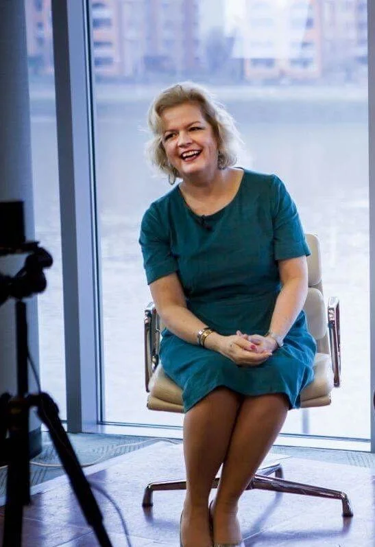 GILL BARHAM  with blonde hair wearing a teal dress, sitting on a chair in front of a large window, smiling during an interview or photoshoot.
