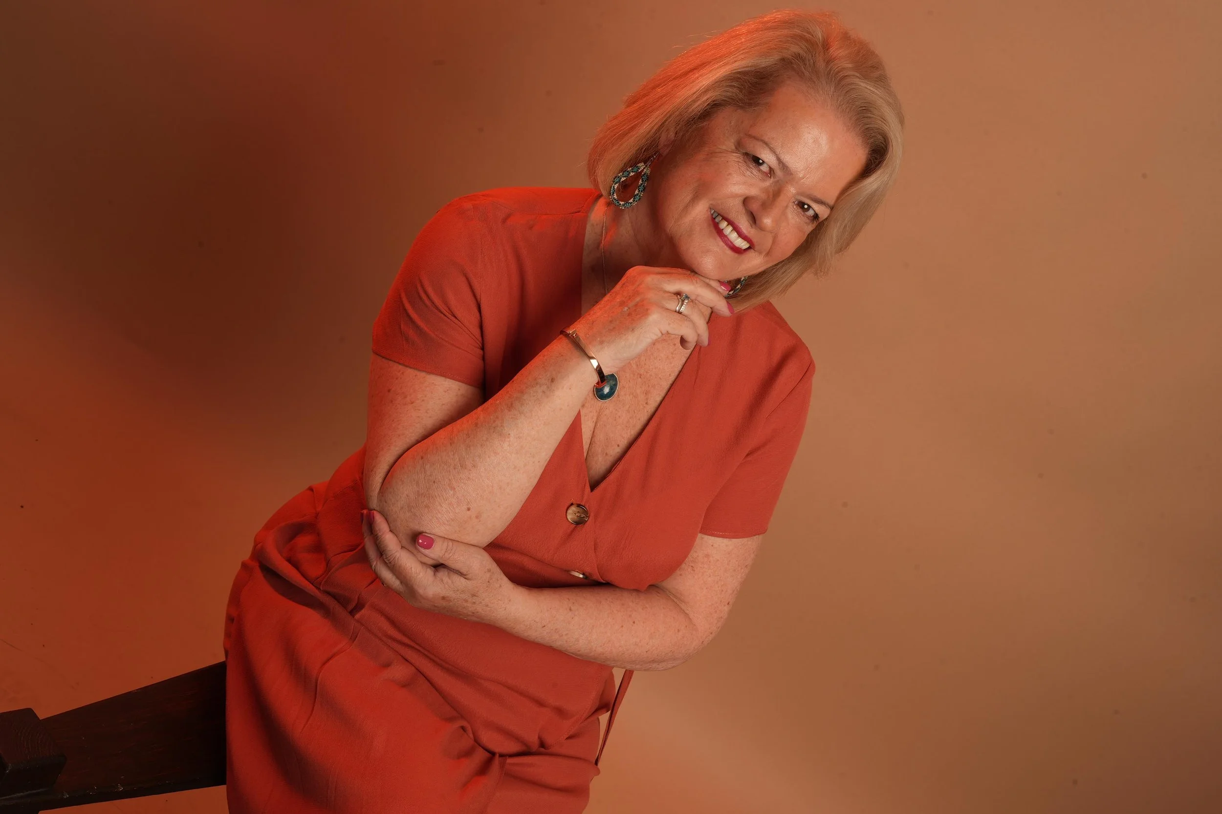 GILL BARHAM with blonde hair, wearing an orange dress, smiling and leaning forward with her hand near her chin, in front of a warm-toned background.