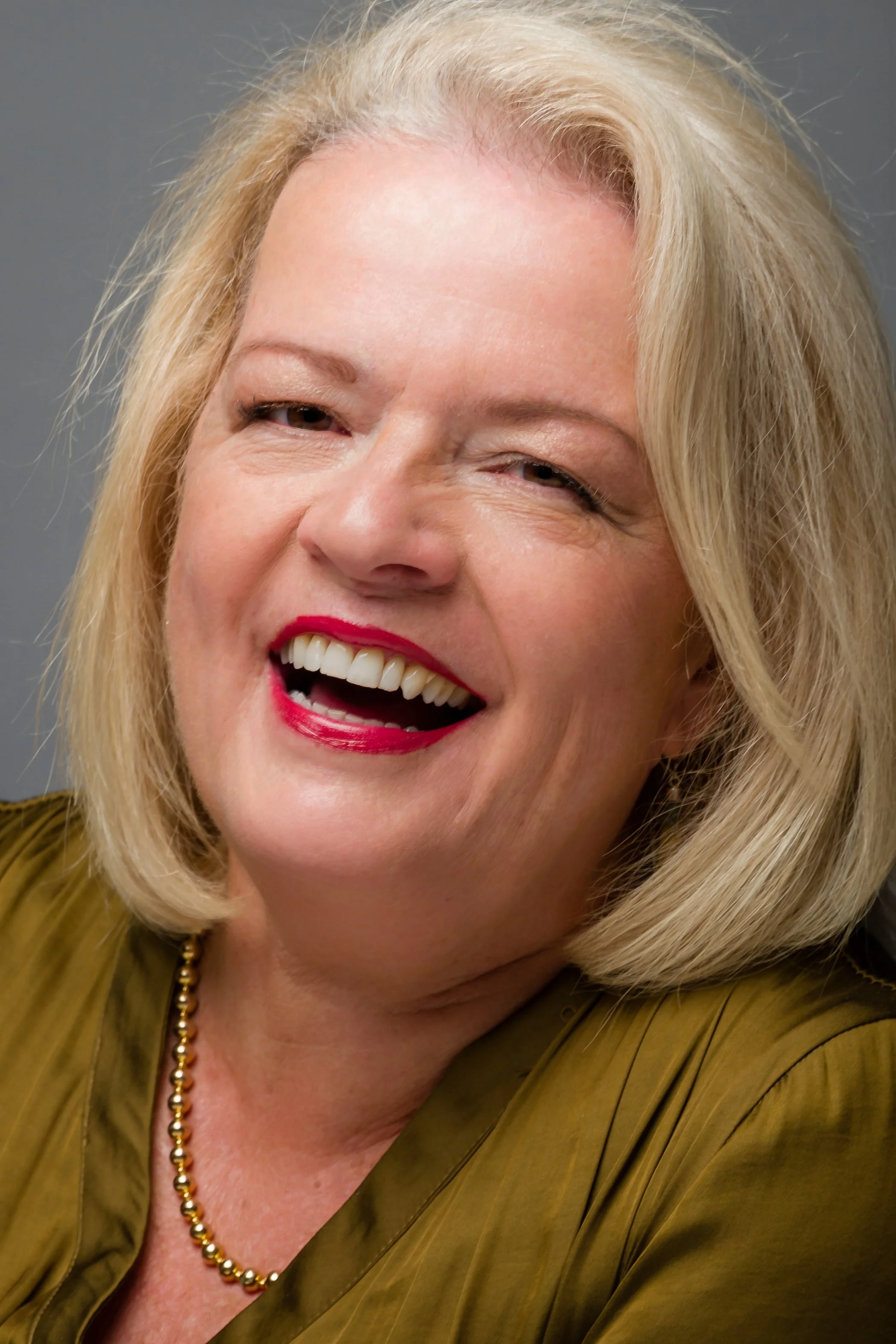 A close-up portrait of a smiling GILL BARHAM with blonde hair, wearing red lipstick, a gold necklace, and a gold top.