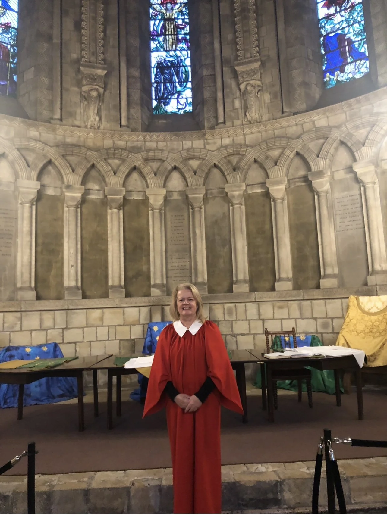 GILL BARHAM dressed in a red choir gown with a white collar standing in a Durham cathedral with ornate stone walls and stained glass windows, behind a table with religious or academic items.