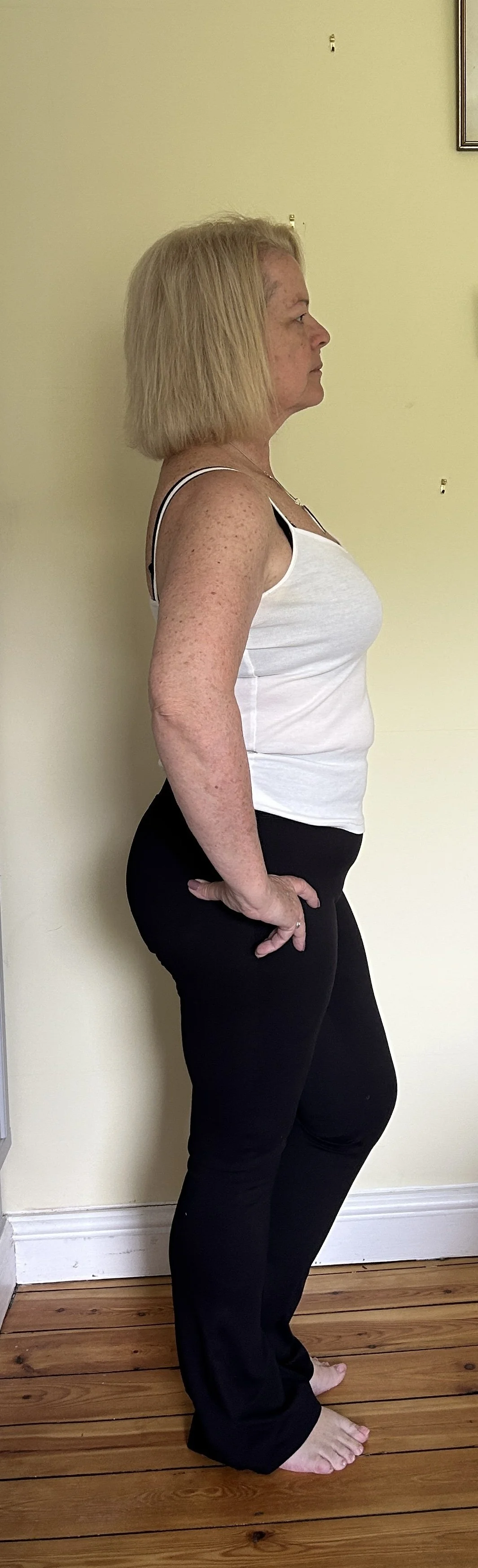 Side view of GILL BARHAM with blonde hair wearing a white tank top and black pants standing barefoot on a wooden floor against a beige wall.