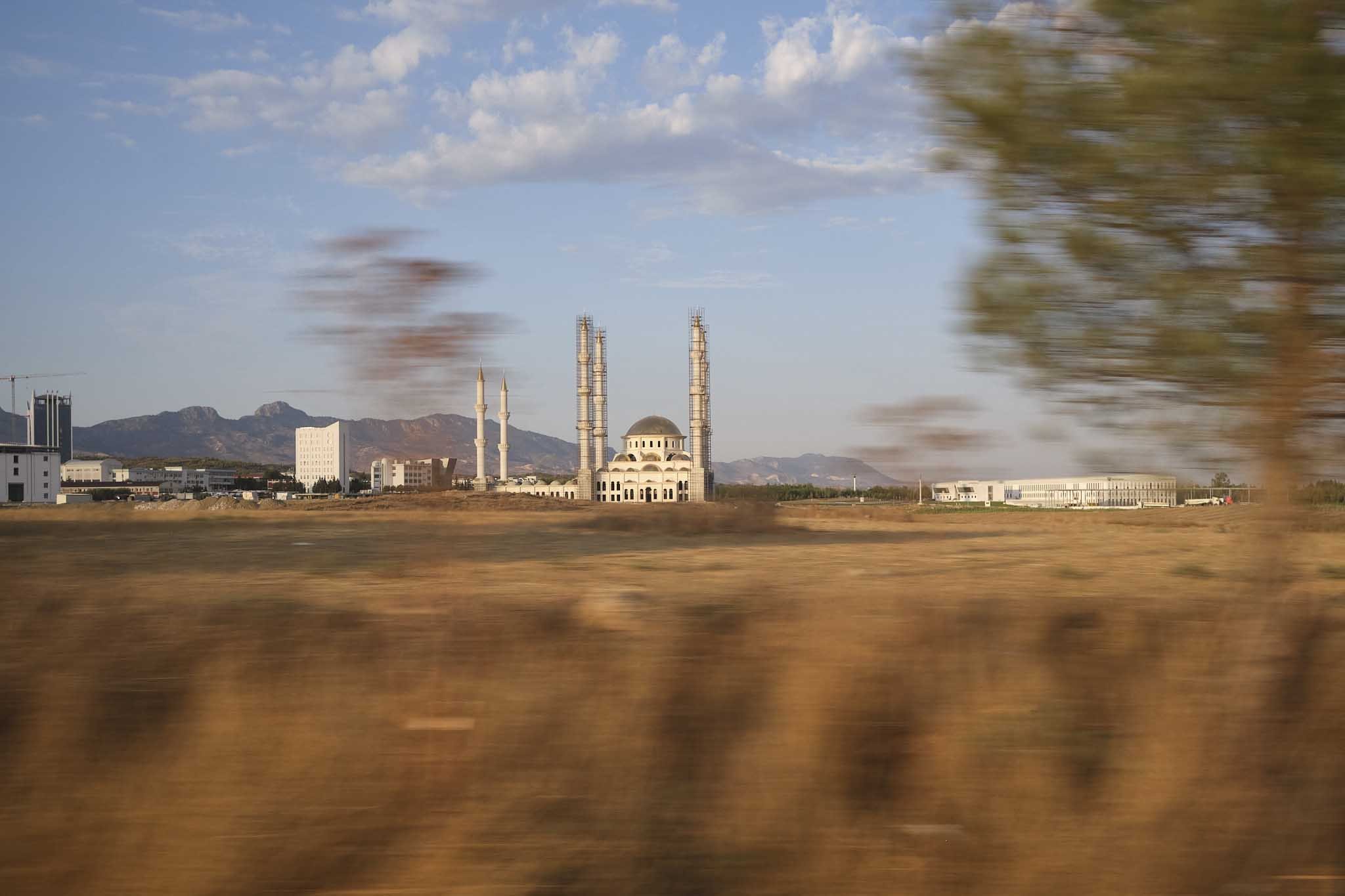 Mosque with minarets and dome in a grassland setting, surrounded by buildings and mountains in the background, slightly blurred foreground trees.