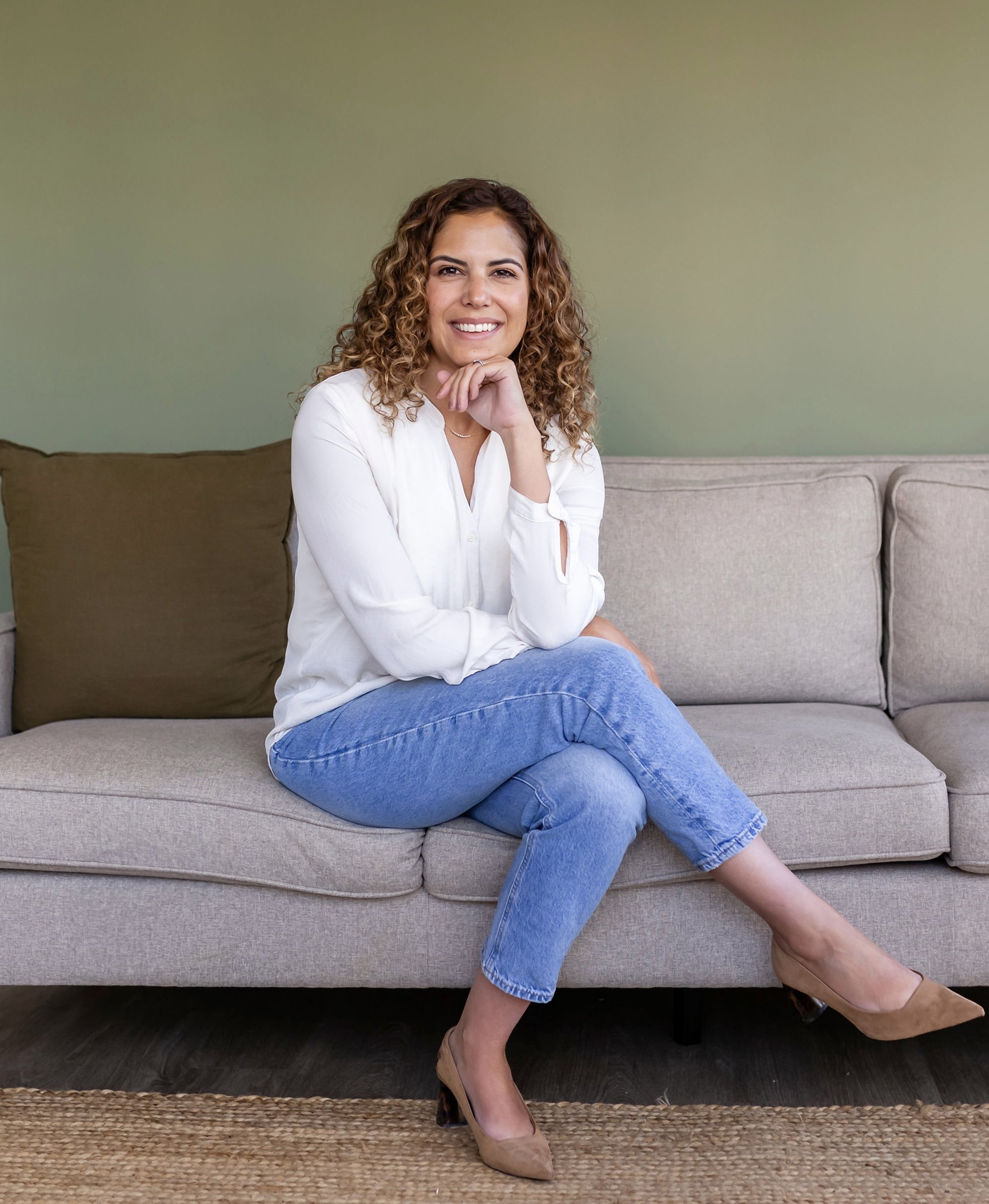 Woman with curly hair sitting on a sofa, wearing a white blouse and jeans, smiling.
