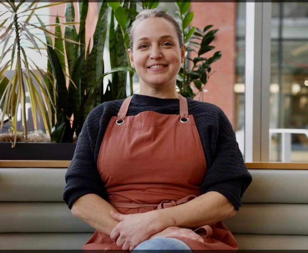 Woman sitting on a bench, wearing an apron, with plants and a glass window in the background.