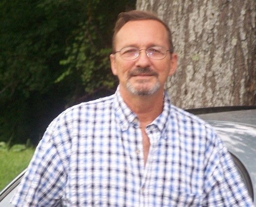 Person in a checkered shirt standing outside near a tree and a car.