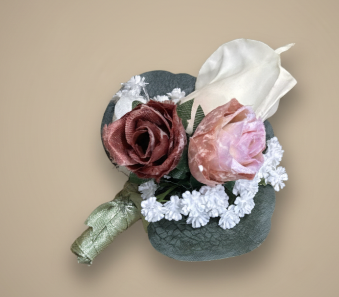 A boutonniere with fabric roses in shades of pink and red, white baby's breath, and a large white leaf, all attached to a dark green stem wrap.