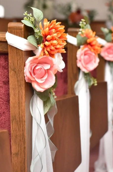 Wooden church pew decorated with pink roses, orange flowers, green leaves, and white ribbons for a wedding.