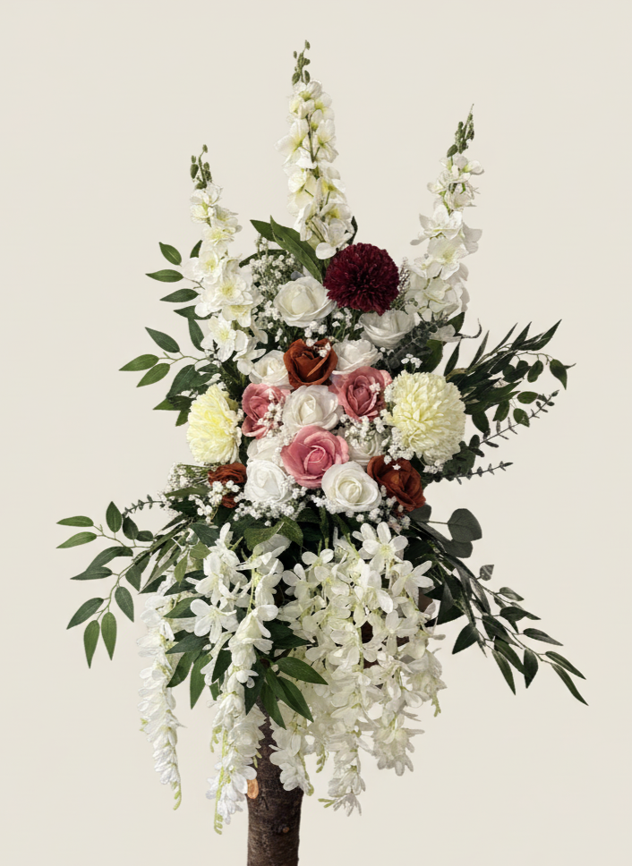A floral arrangement with white, pink, and maroon flowers, including roses, dahlias, and orchids, with green leaves, arranged on a tall stand.