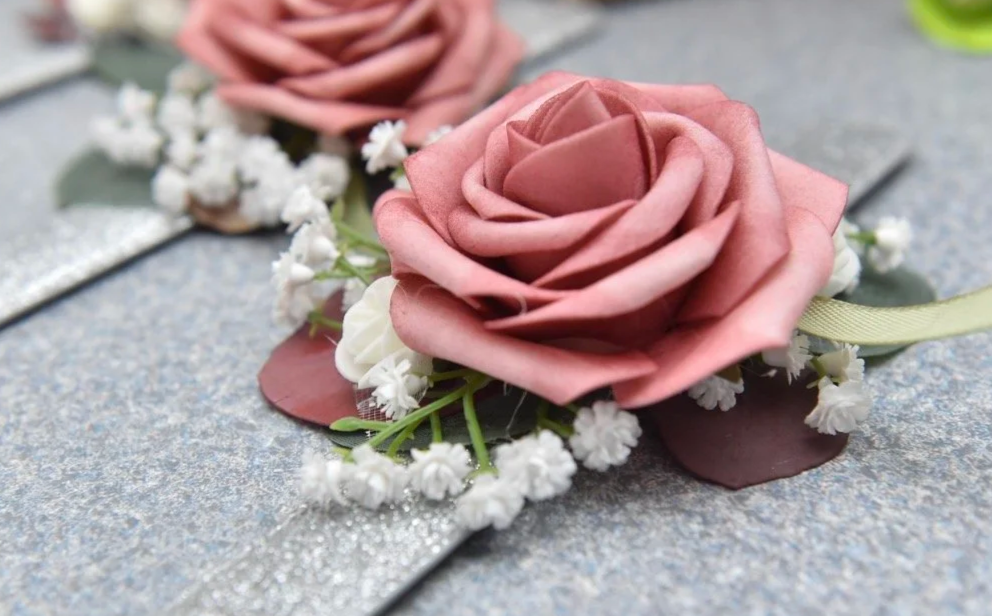 A pink artificial rose with small white baby's breath flowers attached on a gray surface, likely part of a decorative arrangement or memorial.