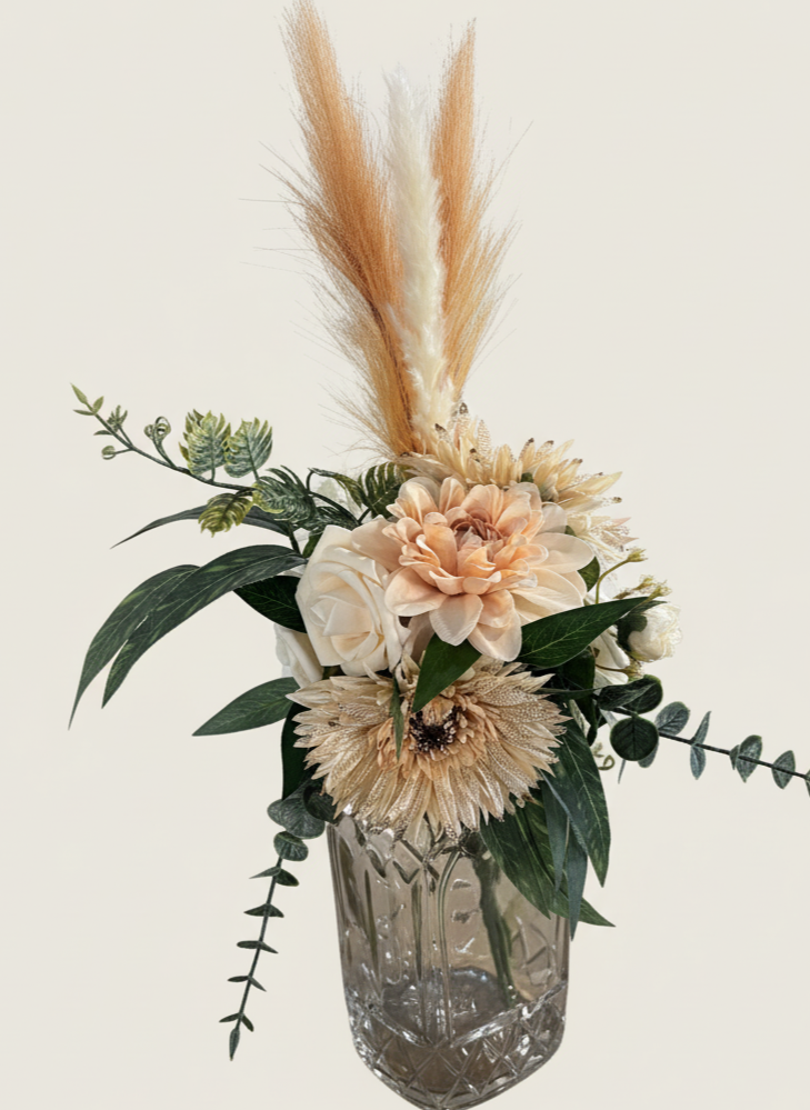 Elegant floral arrangement in a clear glass vase featuring neutral-colored flowers, greenery, and dried pampas grass.
