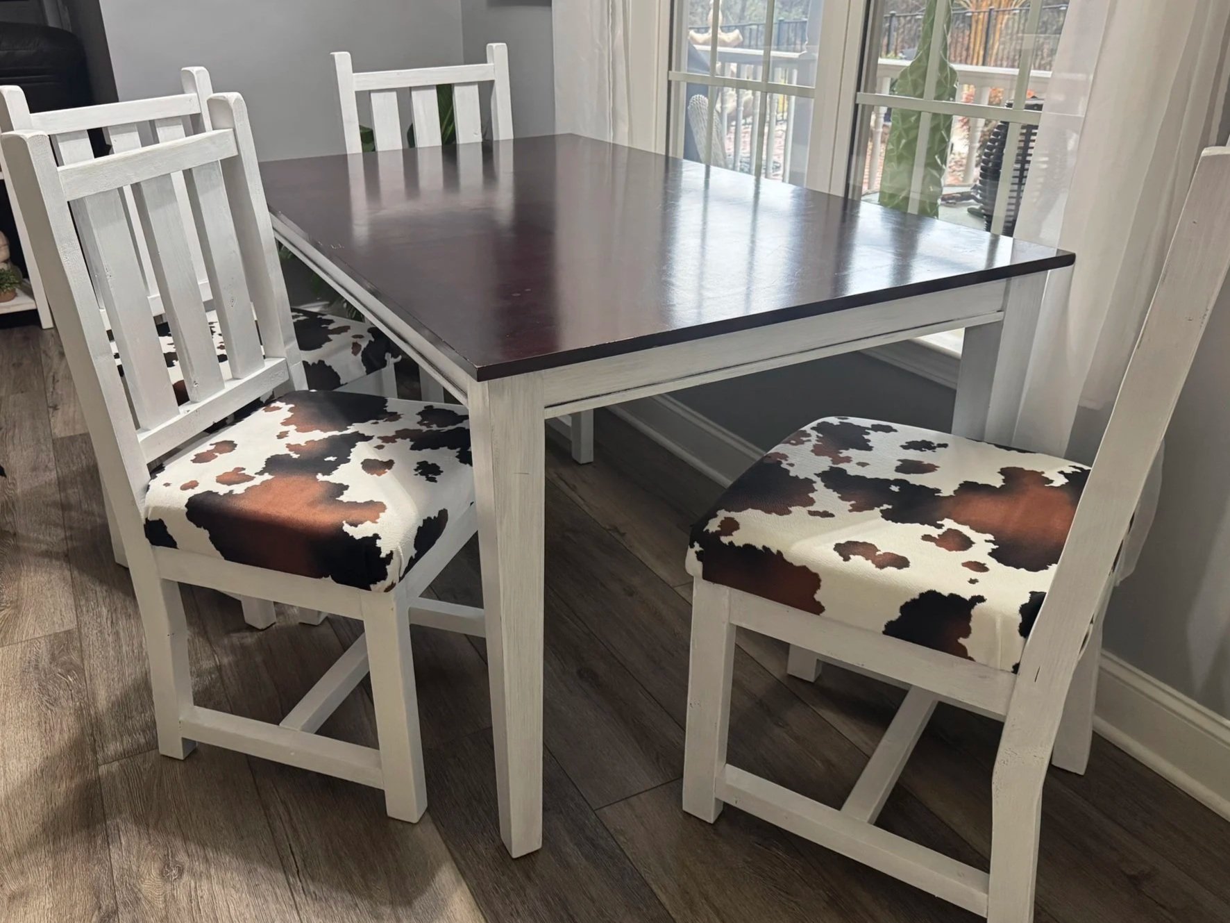 A dining table with four chairs, two with cow print cushions, in a room with wooden floors and large windows.