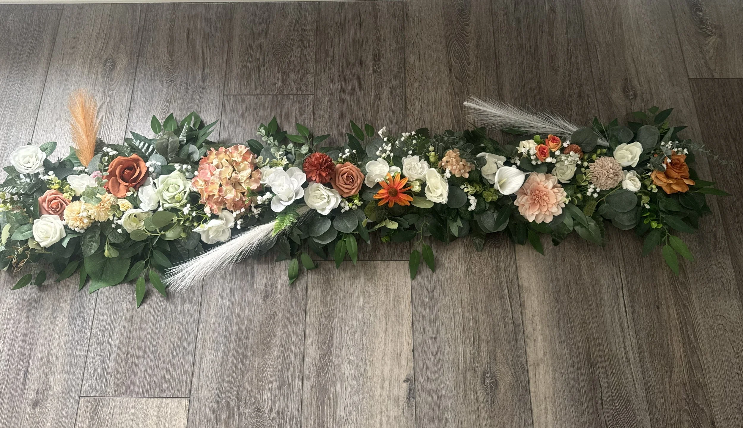 A long floral arrangement with various white, blush, peach, and rust-colored flowers, accented with green foliage and two white pampas grass feathers, placed on a wooden floor.