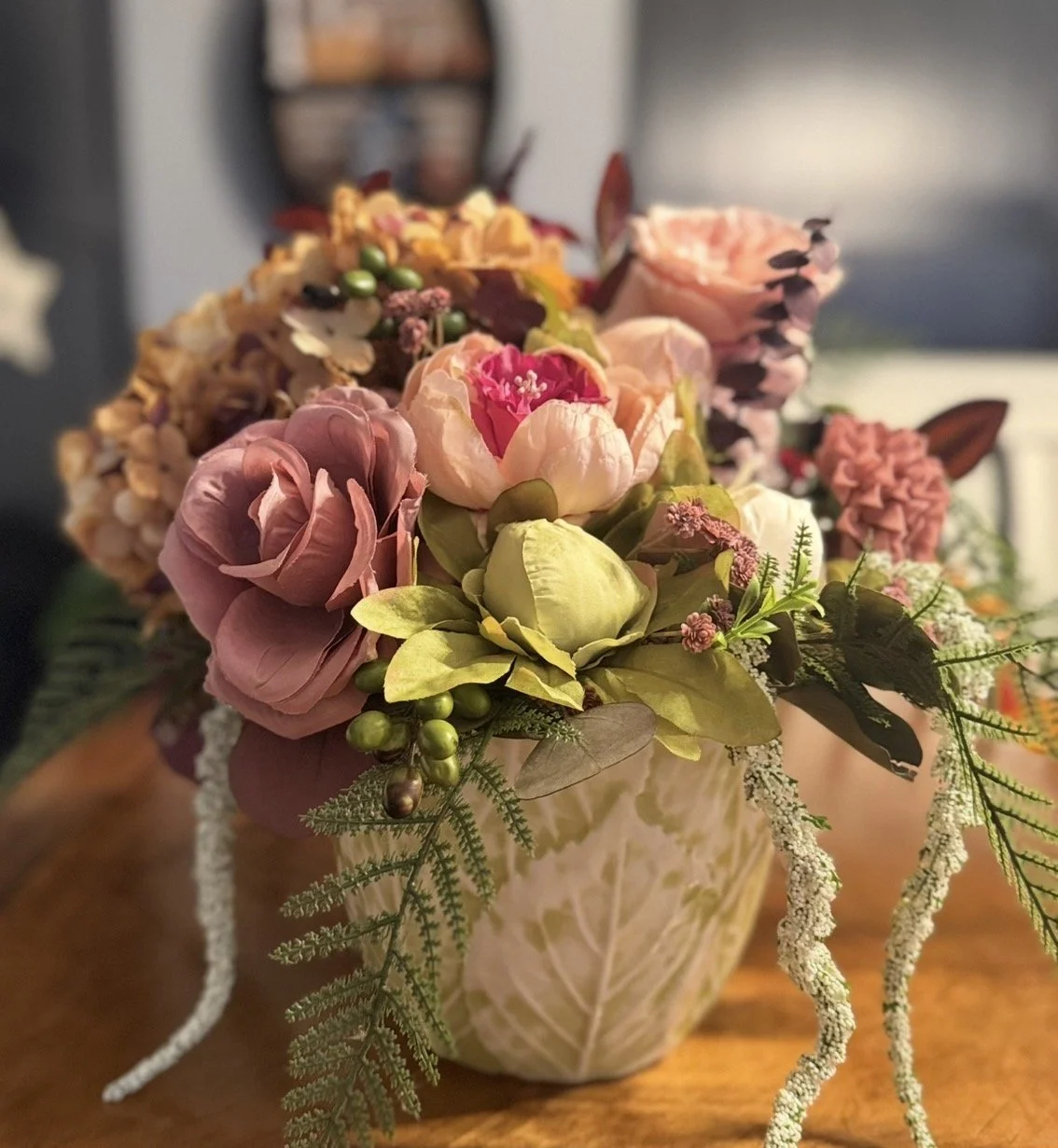 A detailed floral arrangement with pink, cream, and purple flowers in a decorative ceramic vase, placed on a wooden surface.