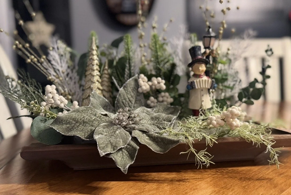 A decorative holiday arrangement with artificial greenery, white berries, frosted leaves, and miniature trees, featuring a small figurine of a boy in a top hat playing a drum, all set in a rectangular wooden tray.