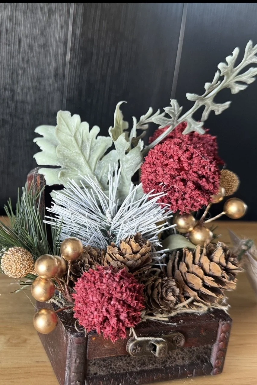 Christmas floral arrangement with pinecones, red flowers, gold berries, and greenery in a wooden box.