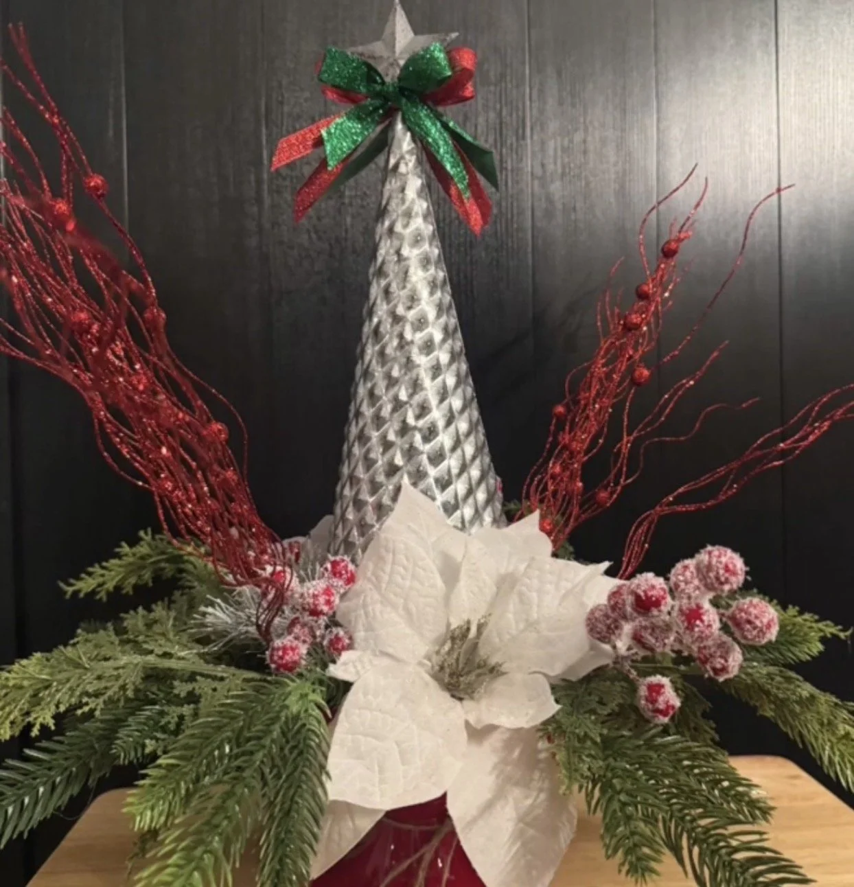 Decorative Christmas arrangement with a silver cone-shaped tree topper, red berries, red twigs, white poinsettia flower, and green pine branches on a wooden surface.